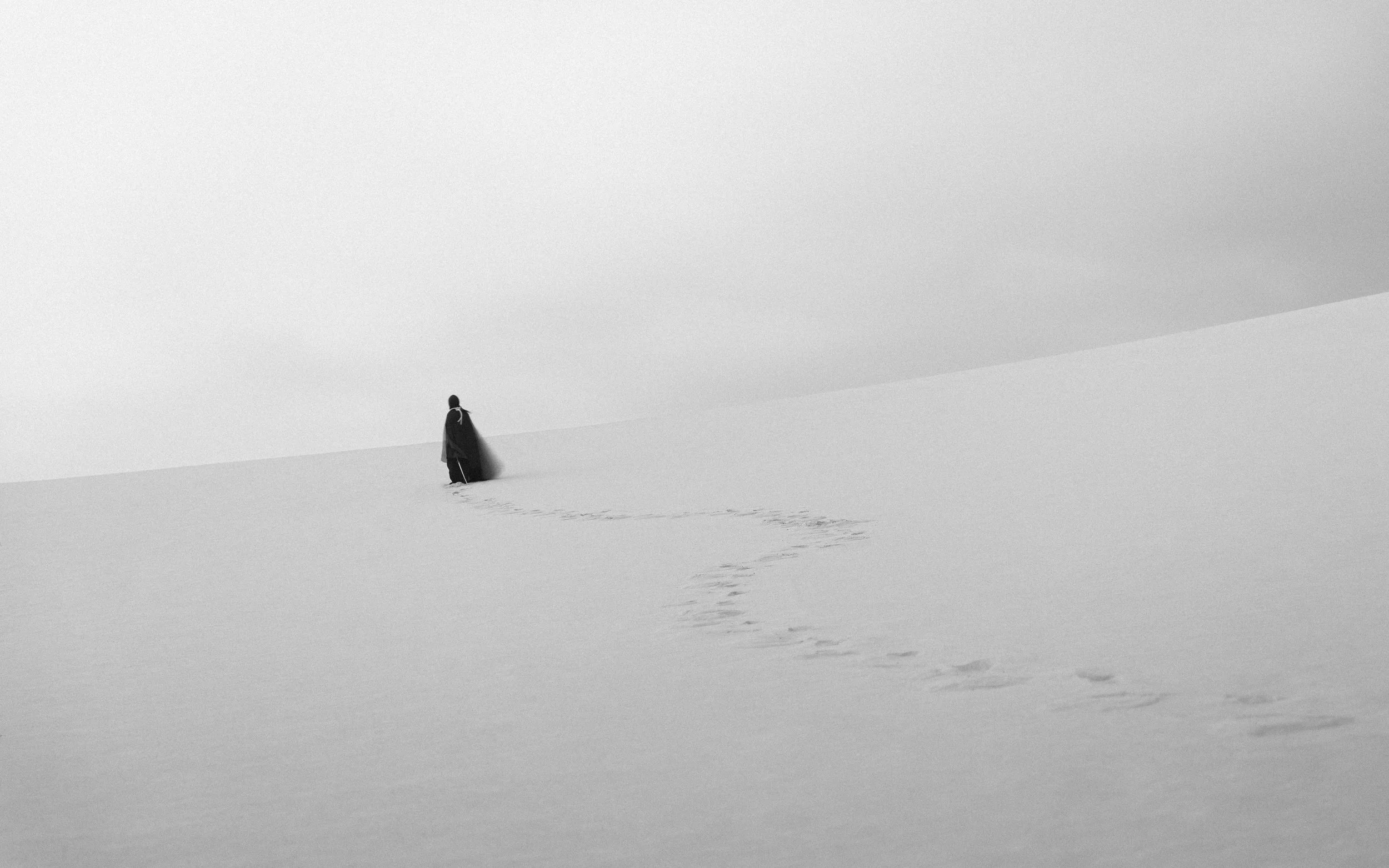 A person walking in a snow-covered or desert landscape, leaving a trail of footprints behind.