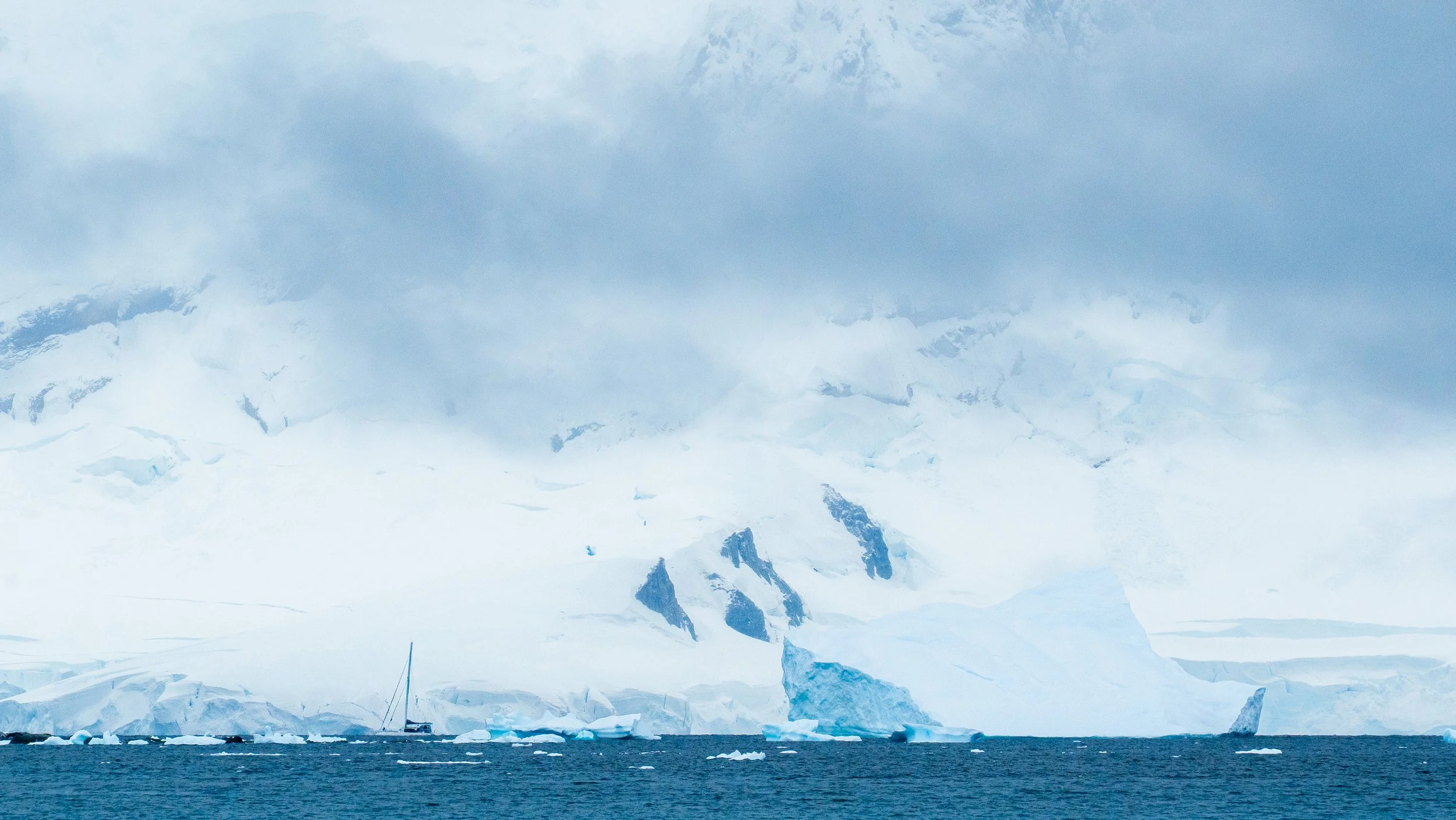 A sailboat near large icebergs in icy blue waters, with snow-covered mountains and clouds in the background.