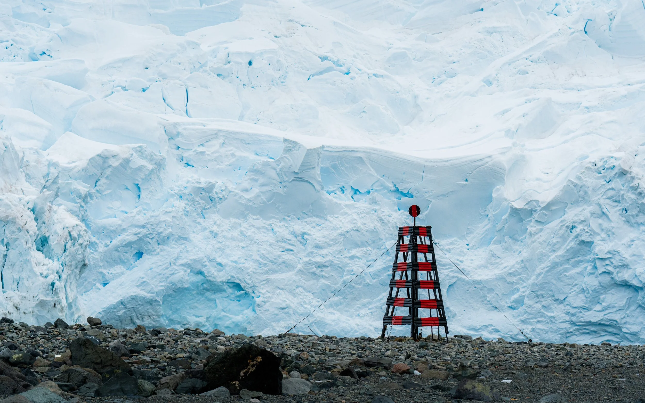 Large icy glacier with a red and black beacon or marker at the bottom in a rocky landscape.