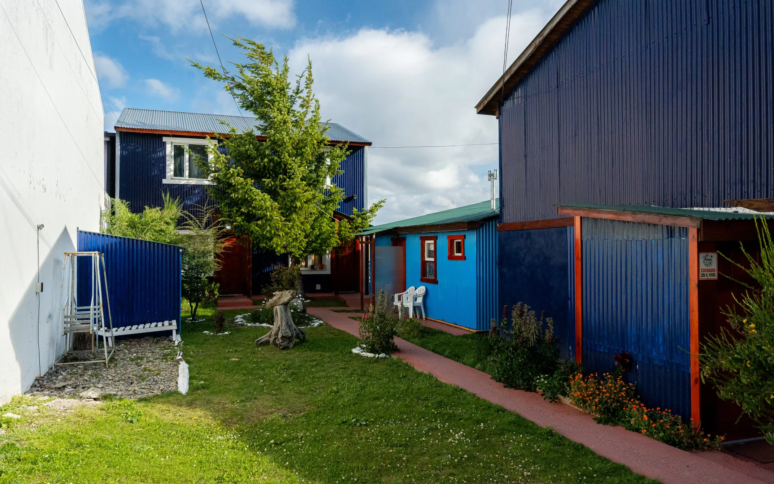 A backyard with a green lawn, garden beds, and a tree trunk, surrounded by colorful houses with blue, red, and white walls and metal roofs, under a partly cloudy sky.
