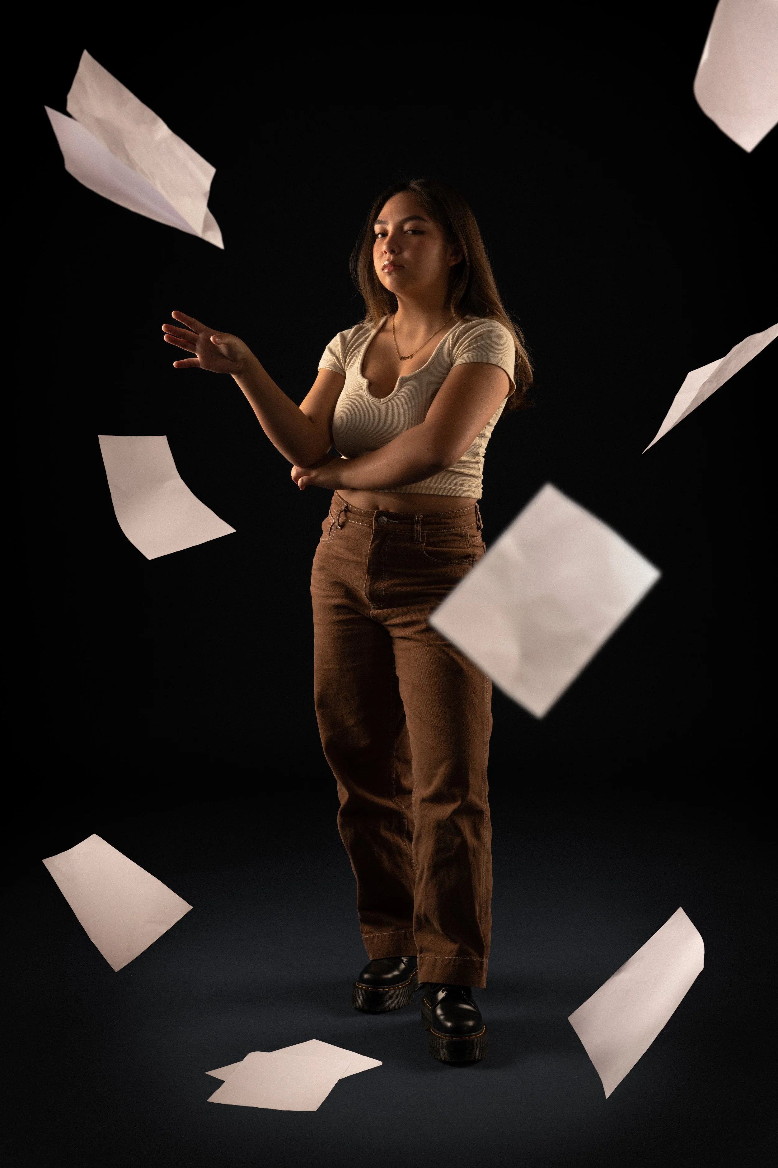Young woman with long brown hair, wearing a beige crop top and brown pants, standing against a black background with scattered white sheets of paper floating around her.