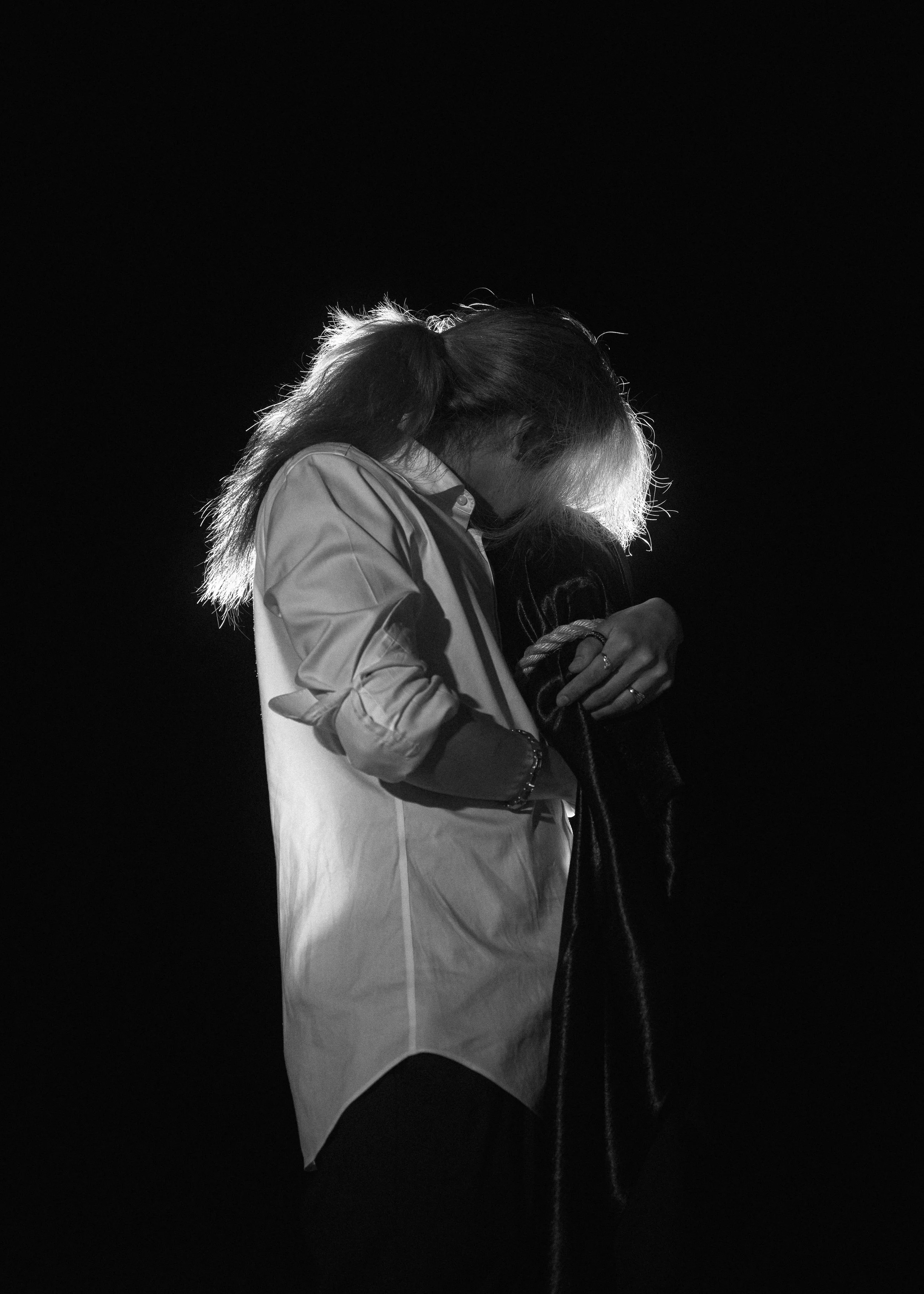 Black and white photo of a person with long hair, wearing a button-up shirt, hugging or holding a dog, against a dark background.