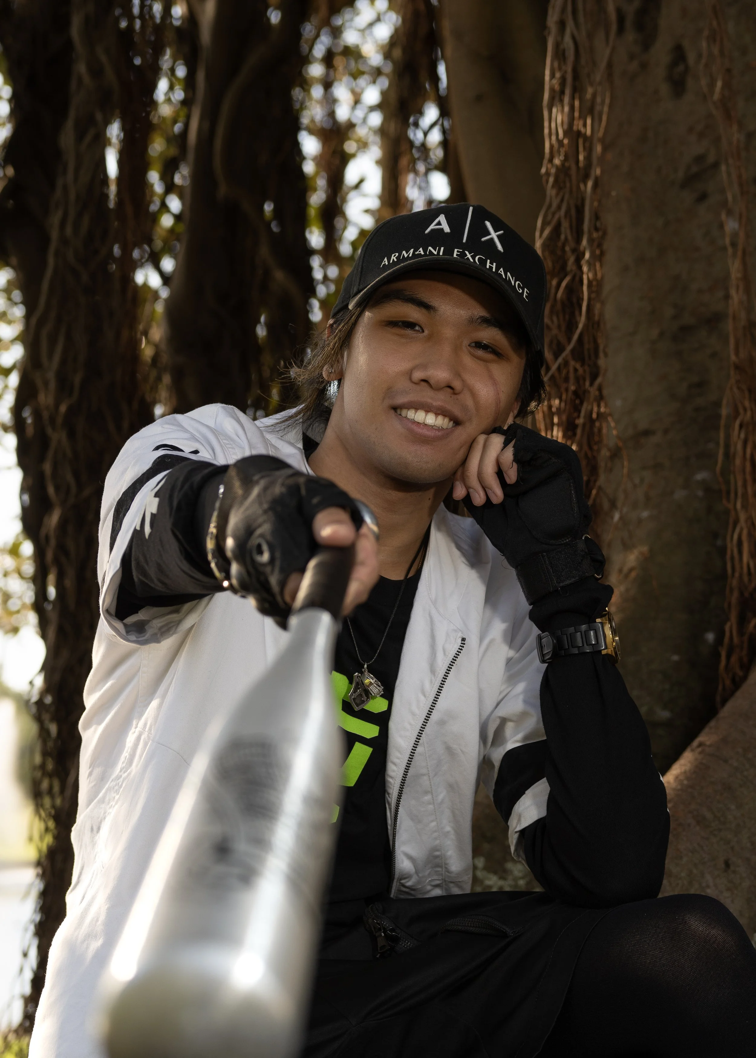 Young man with black hair and a smile, wearing a black cap with white text and a white and black jacket, taking a selfie outdoors against a background of tree trunks and sunlight filtering through leaves.