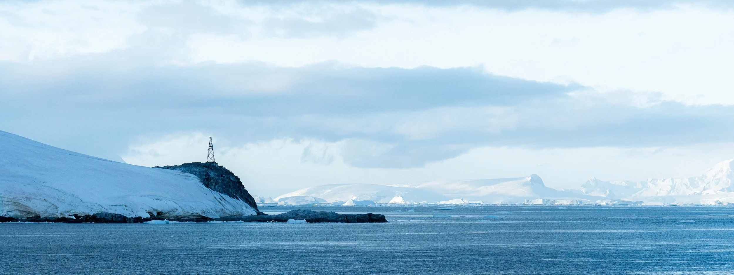Arctic landscape with icy shoreline, snow-covered mountains, and a communication tower on a rocky outcrop.