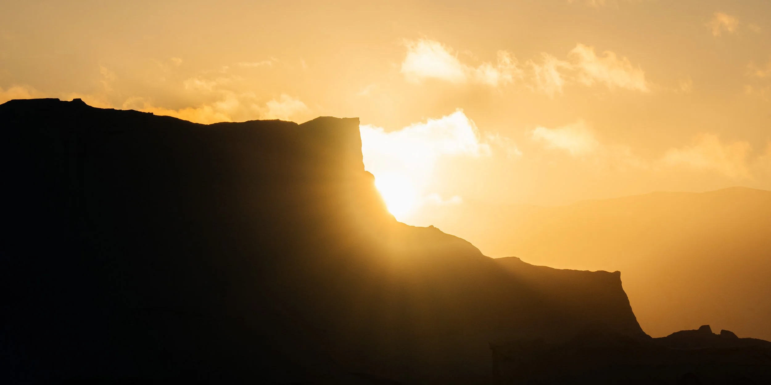 Sun setting behind silhouette of mountain or cliff with clouds in the sky
