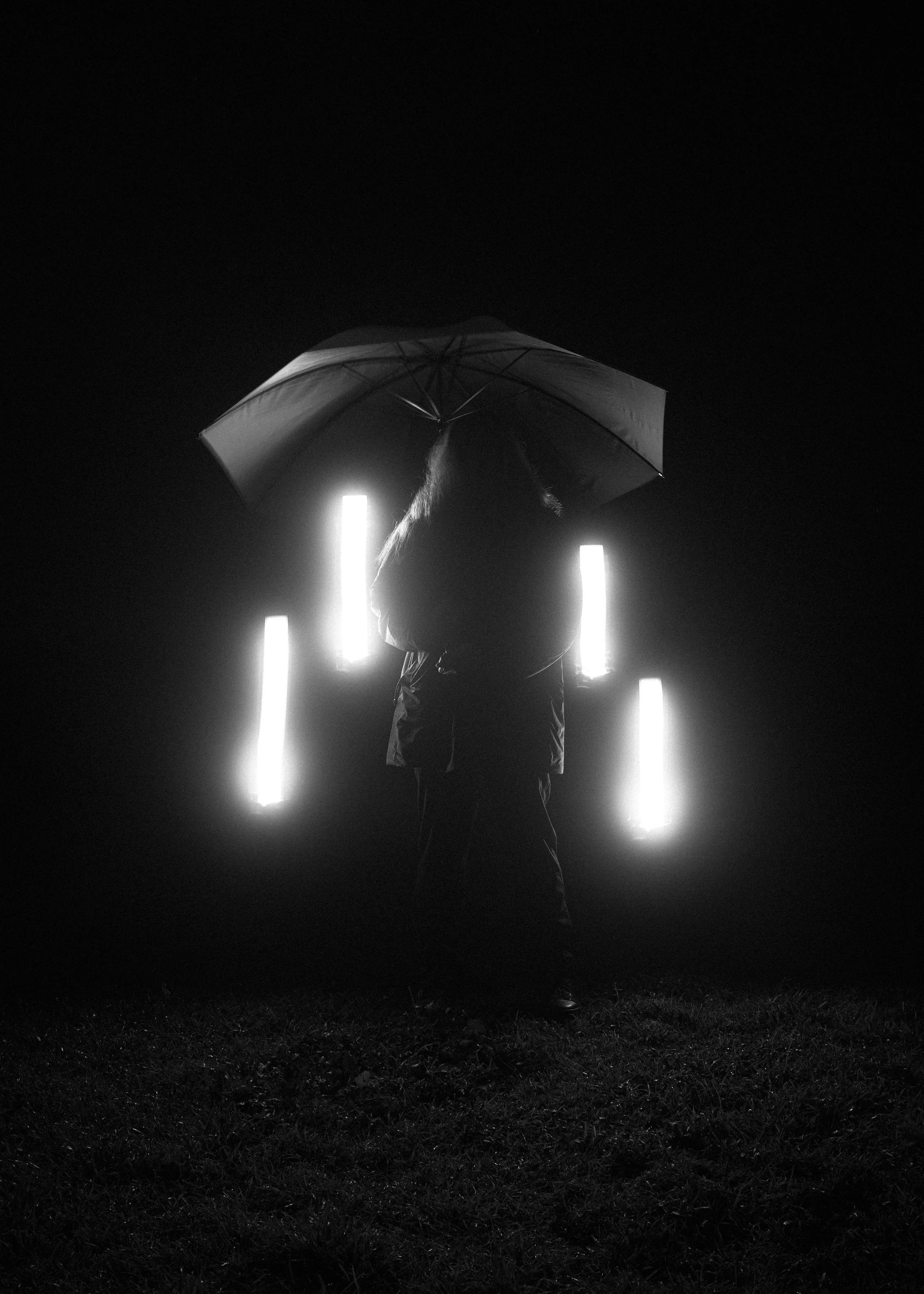 Person holding an umbrella standing in front of bright vertical lights in a dark outdoor setting at night.