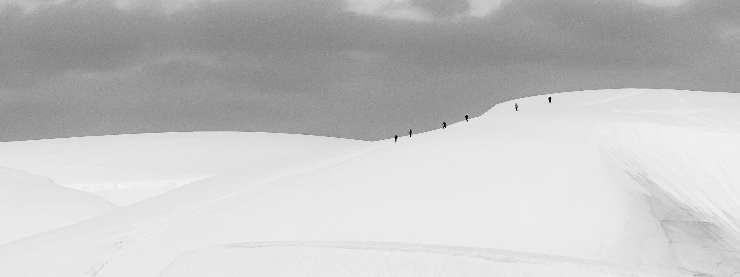 Snow-covered landscape with seven hikers walking in a line along a ridge under a cloudy sky.