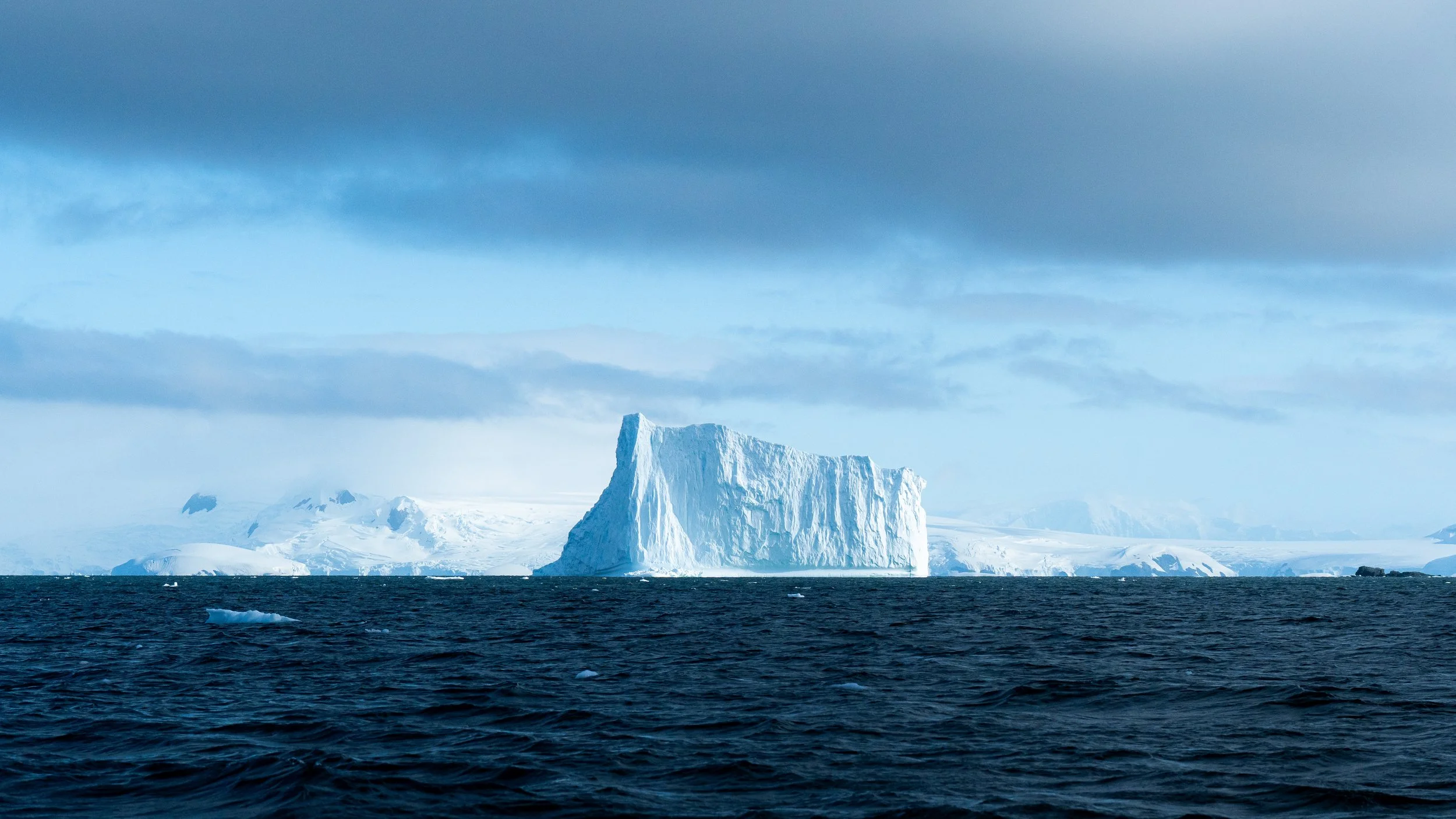 A large iceberg floating in the ocean with a cloudy sky overhead.