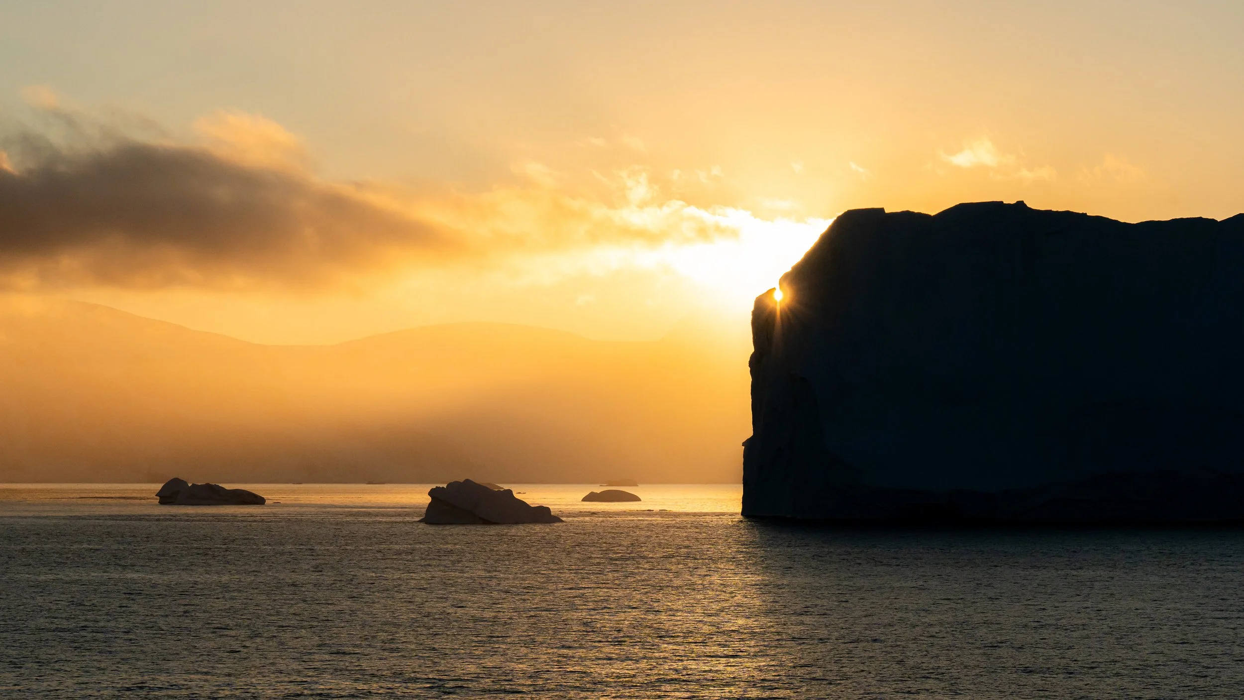 Sunset over the ocean with large dark rock formations in the water and a cliff on the right side, with the sun partially hidden behind it.