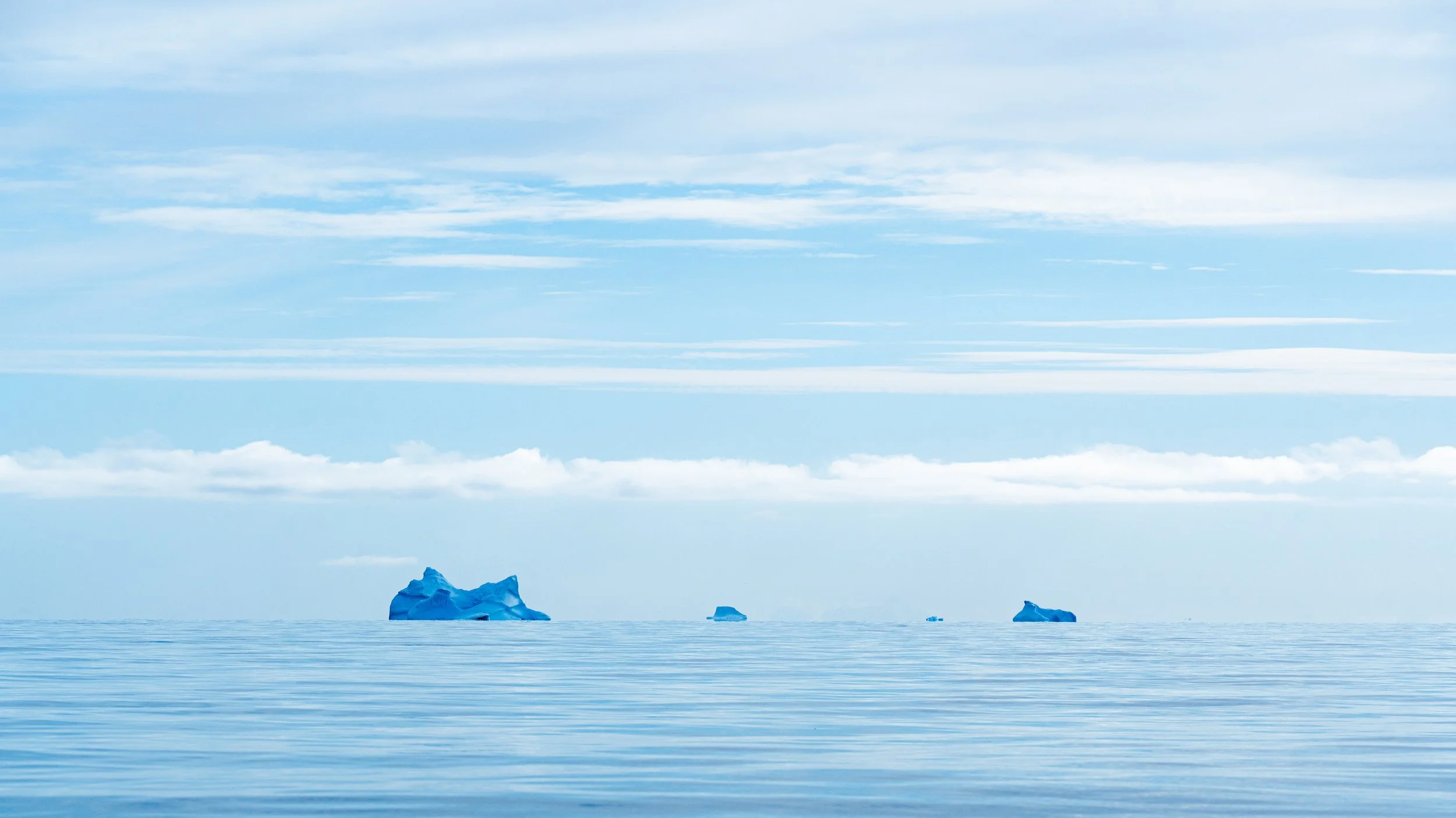 A vast icy ocean with several icebergs floating on the water under a blue sky with high-altitude clouds.