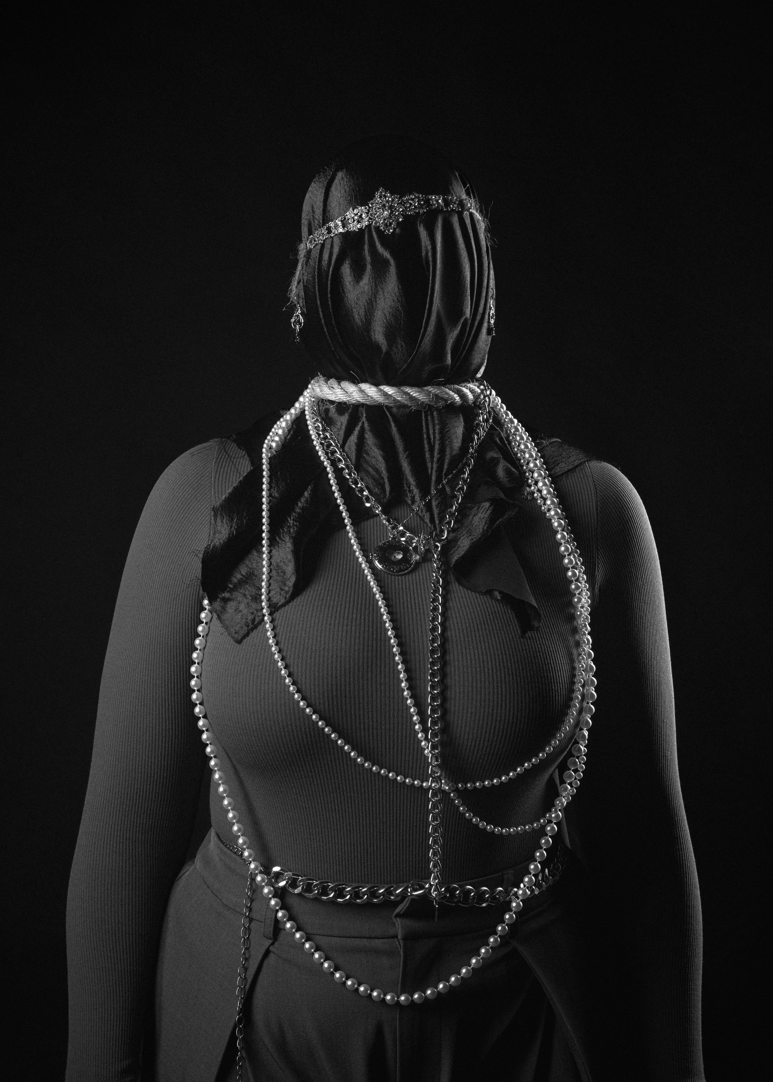Black and white photo of a woman with jewelry and a headscarf, viewed from the back against a dark background.