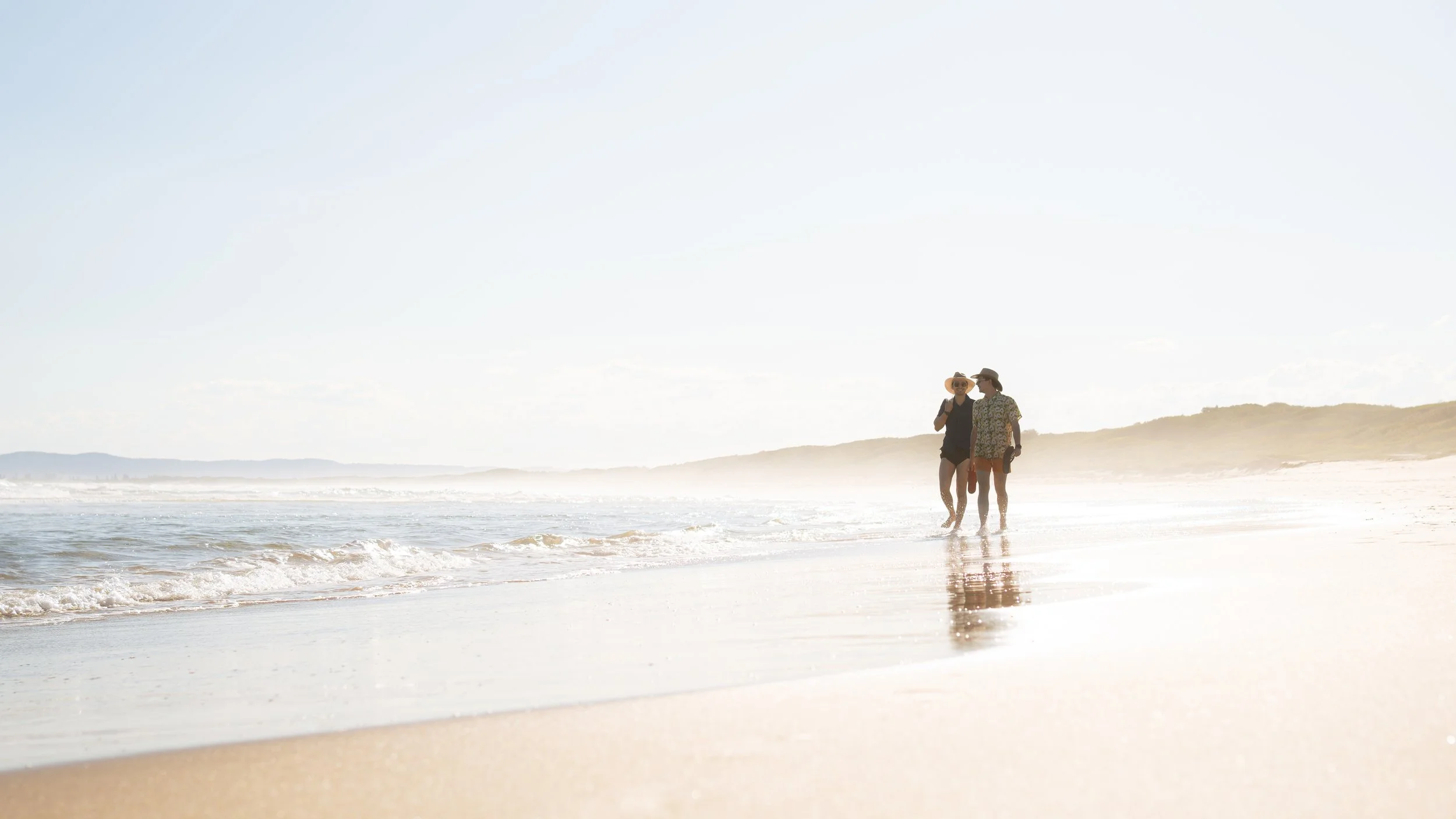 Two women wearing sun hats walking along the shoreline of a beach with calm waves and a distant landmass under a clear sky.