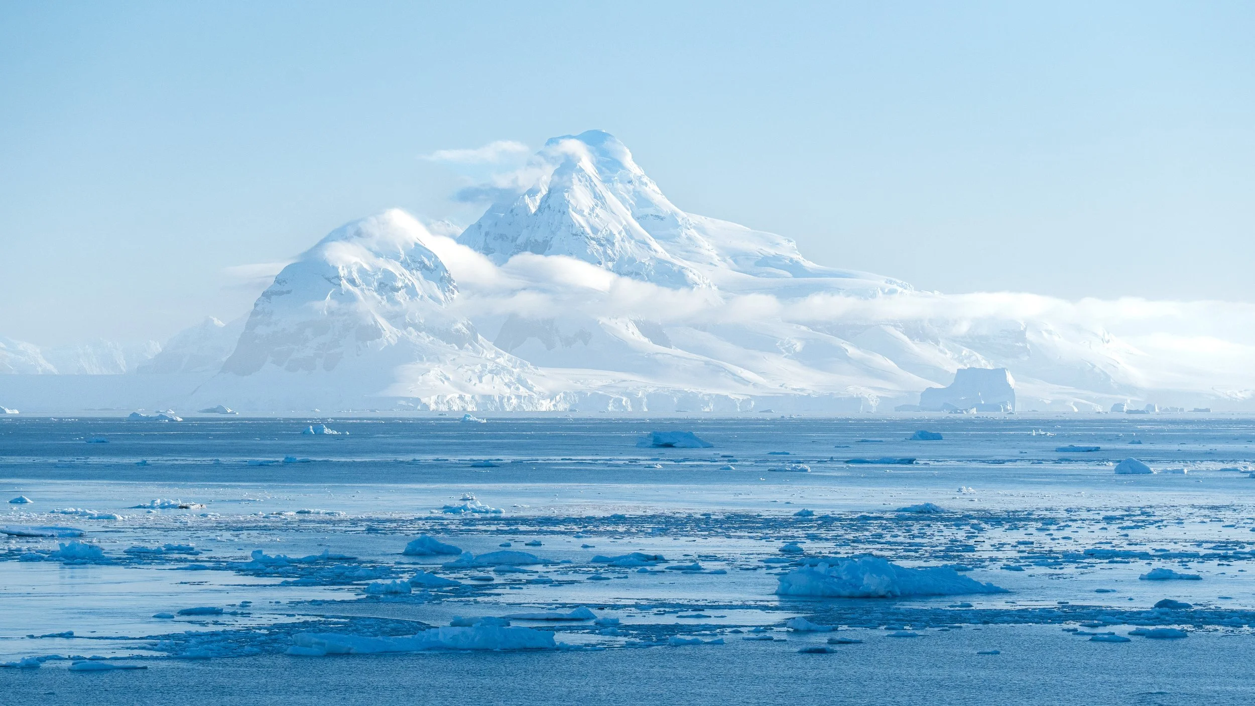 A vast icy landscape with snow-covered mountains, some clouds near the peaks, and floating ice and snow on the water's surface.