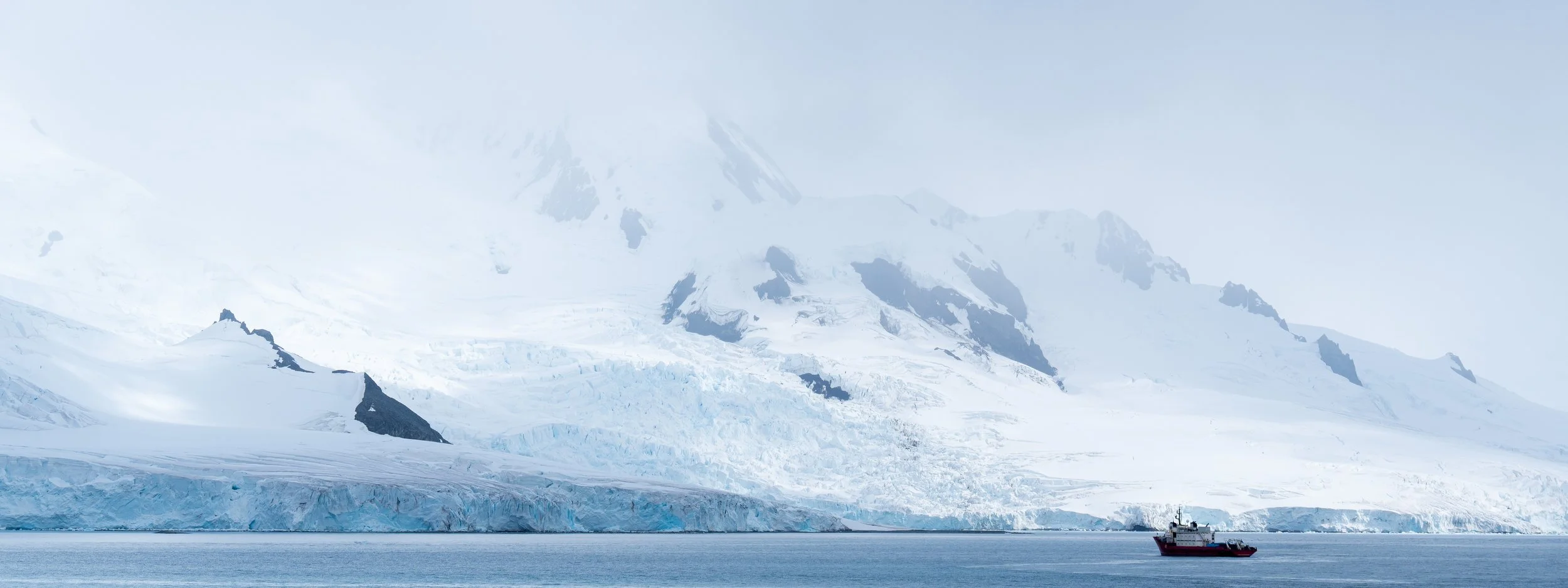 A red ship sailing in icy waters near a glacier with snow-covered mountains in the background.