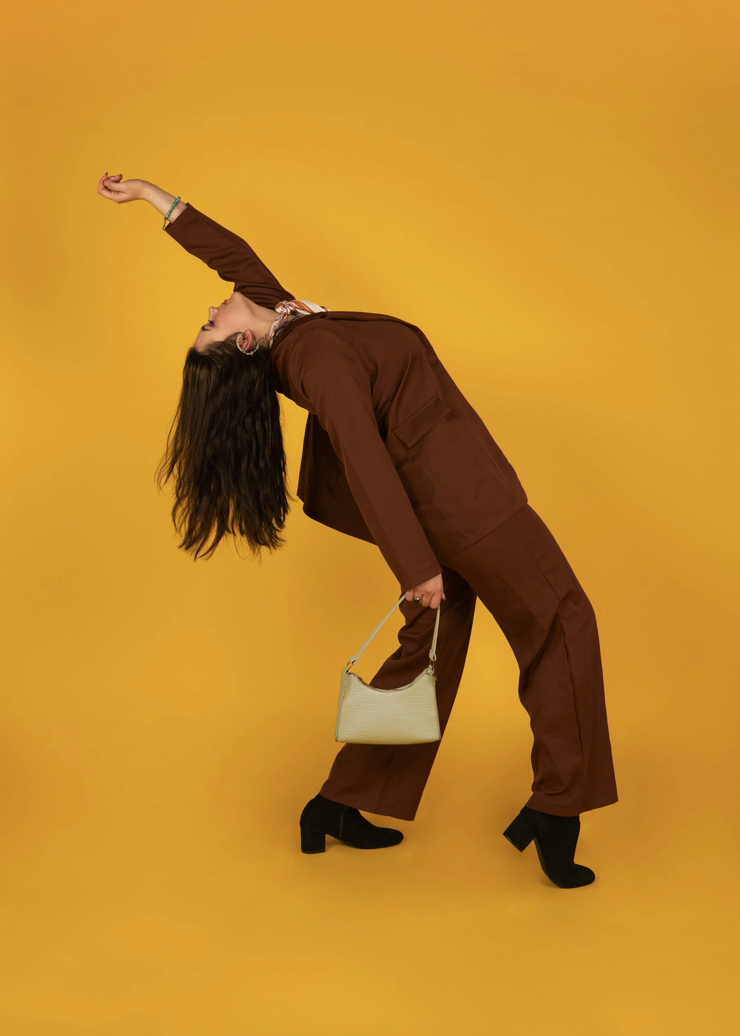 A woman in a brown suit and black heeled boots is posing against a yellow background, bending backward with her left arm extended upward and her right hand holding a white handbag.