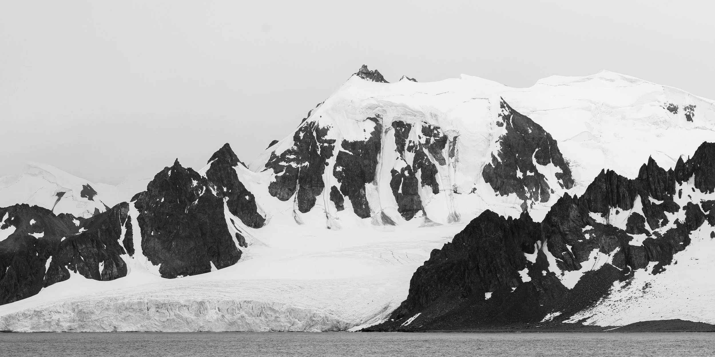 Black and white photograph of a snowy mountain range with dark rocky peaks in the foreground and a body of water at the base.