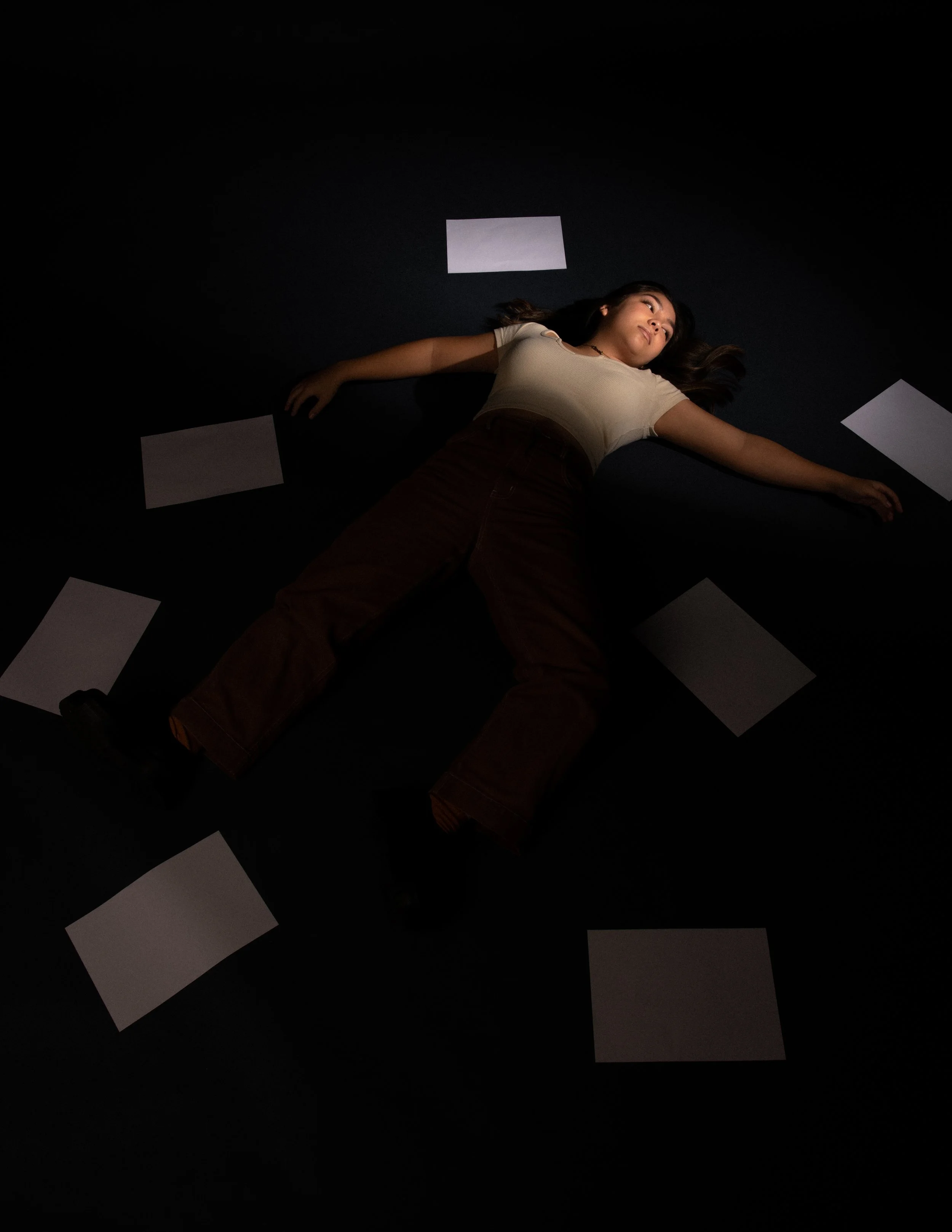 A woman lying on a dark floor surrounded by blank sheets of paper.