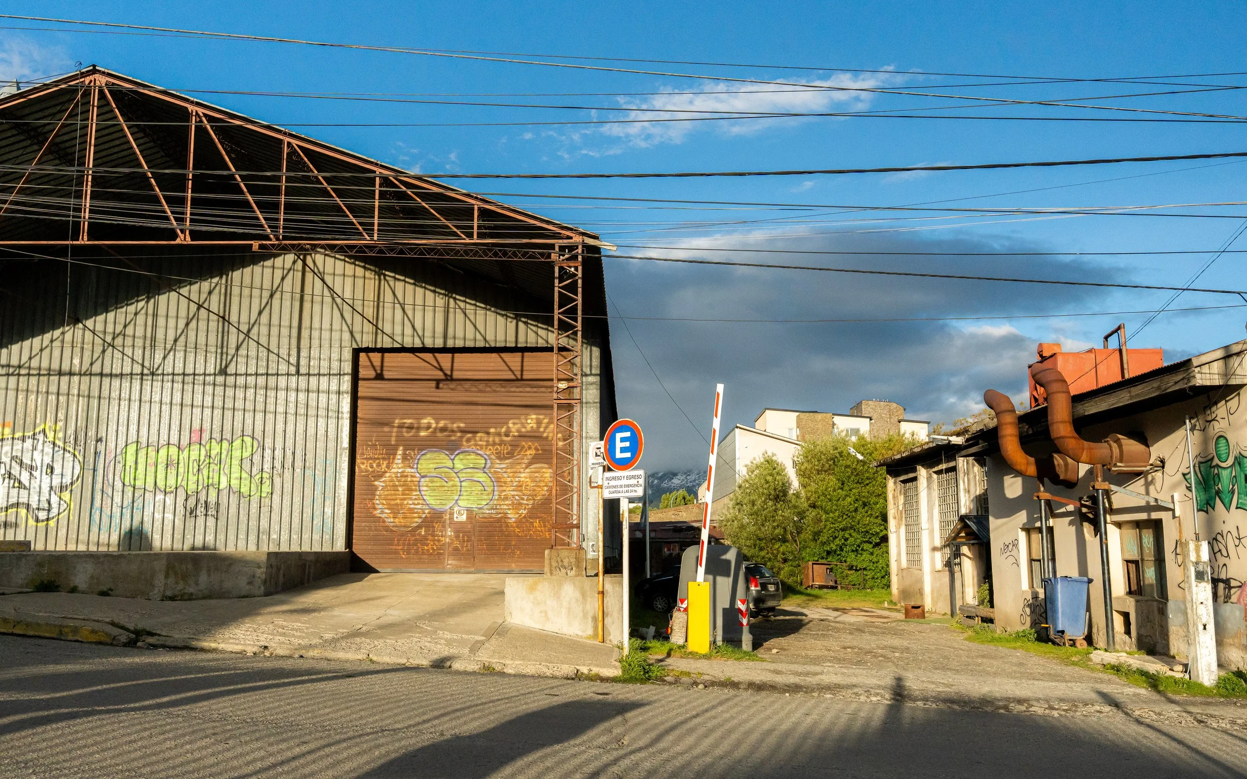 An industrial building with graffiti on a metal wall, a brown door, and overhead wires against a blue sky with clouds.