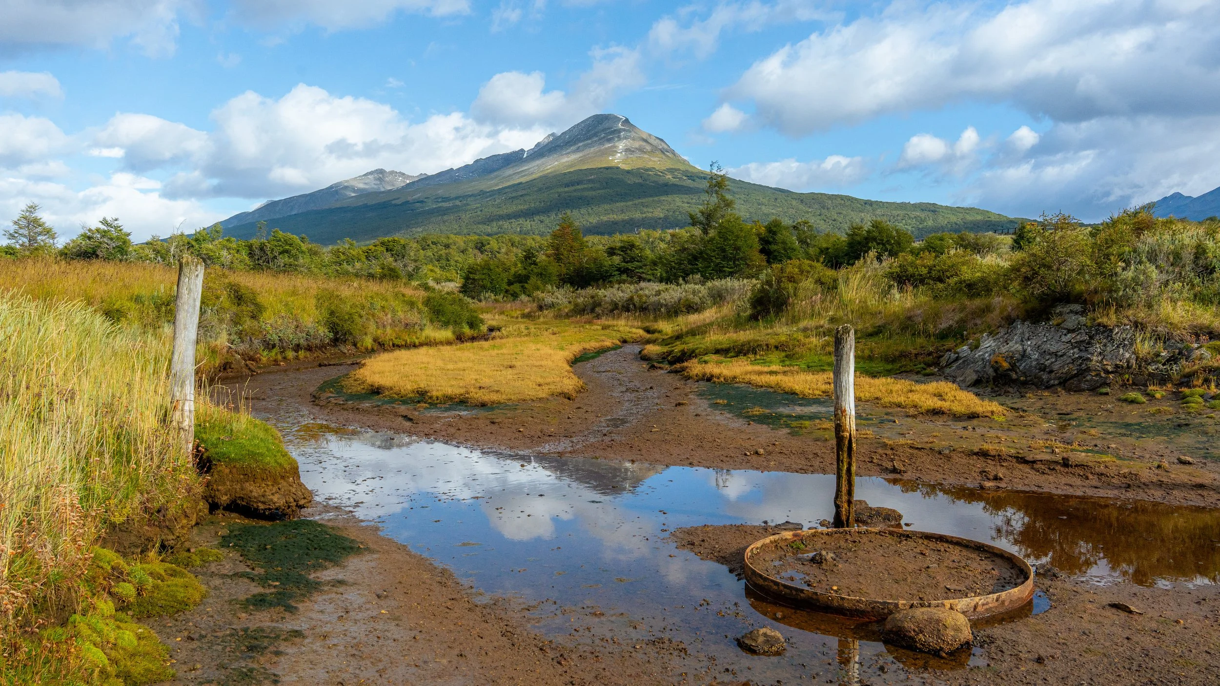 Mountain with a cloud-covered peak, green hillside, and a muddy path with puddles in a rural landscape.