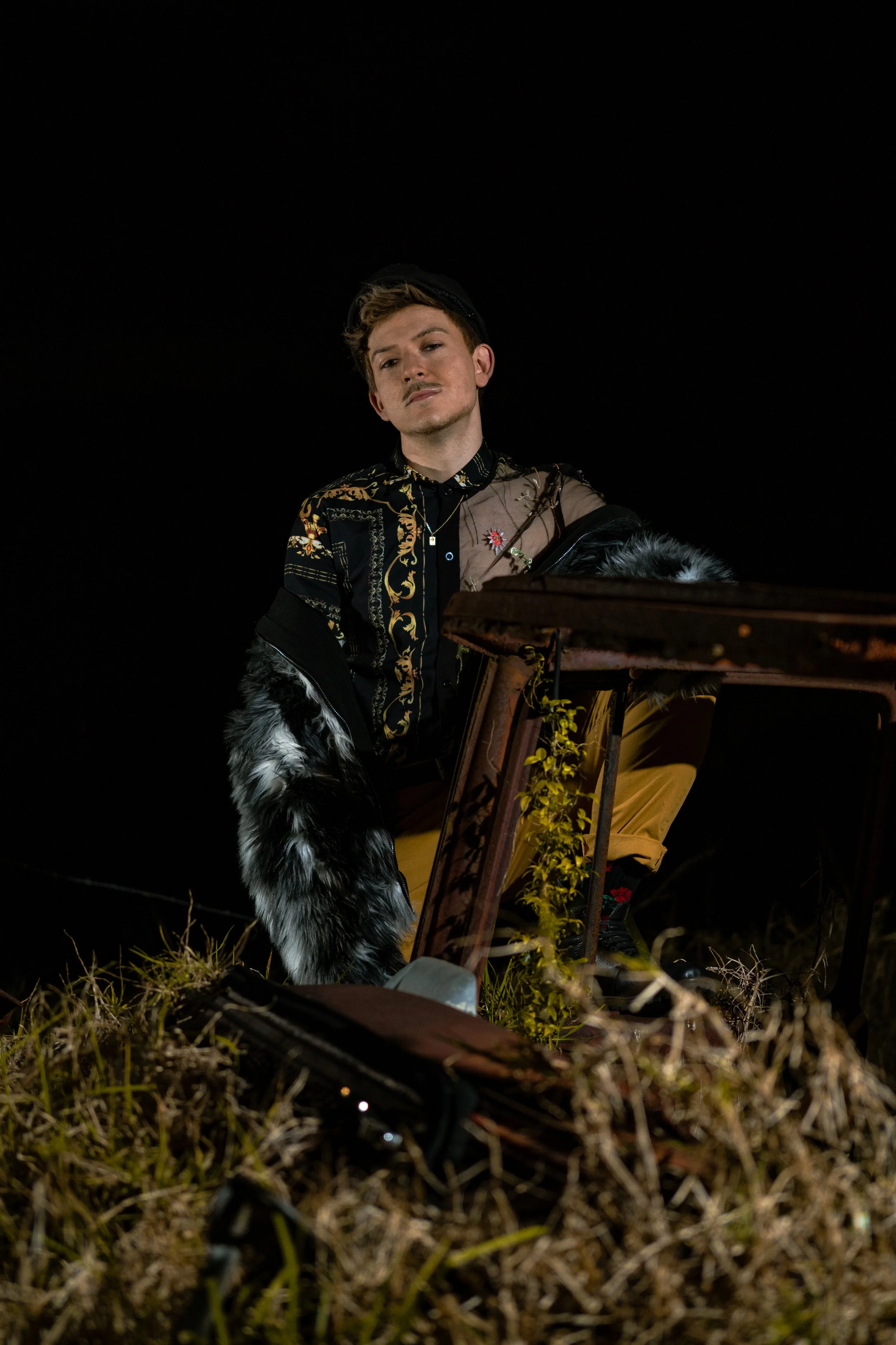 Young man with light skin and short hair seated outdoors at night, wearing a black patterned shirt, dark jacket with furry cuffs, and a necklace, with a black cap tilted backwards, leaning on a wooden ladder with yellow plants growing around.