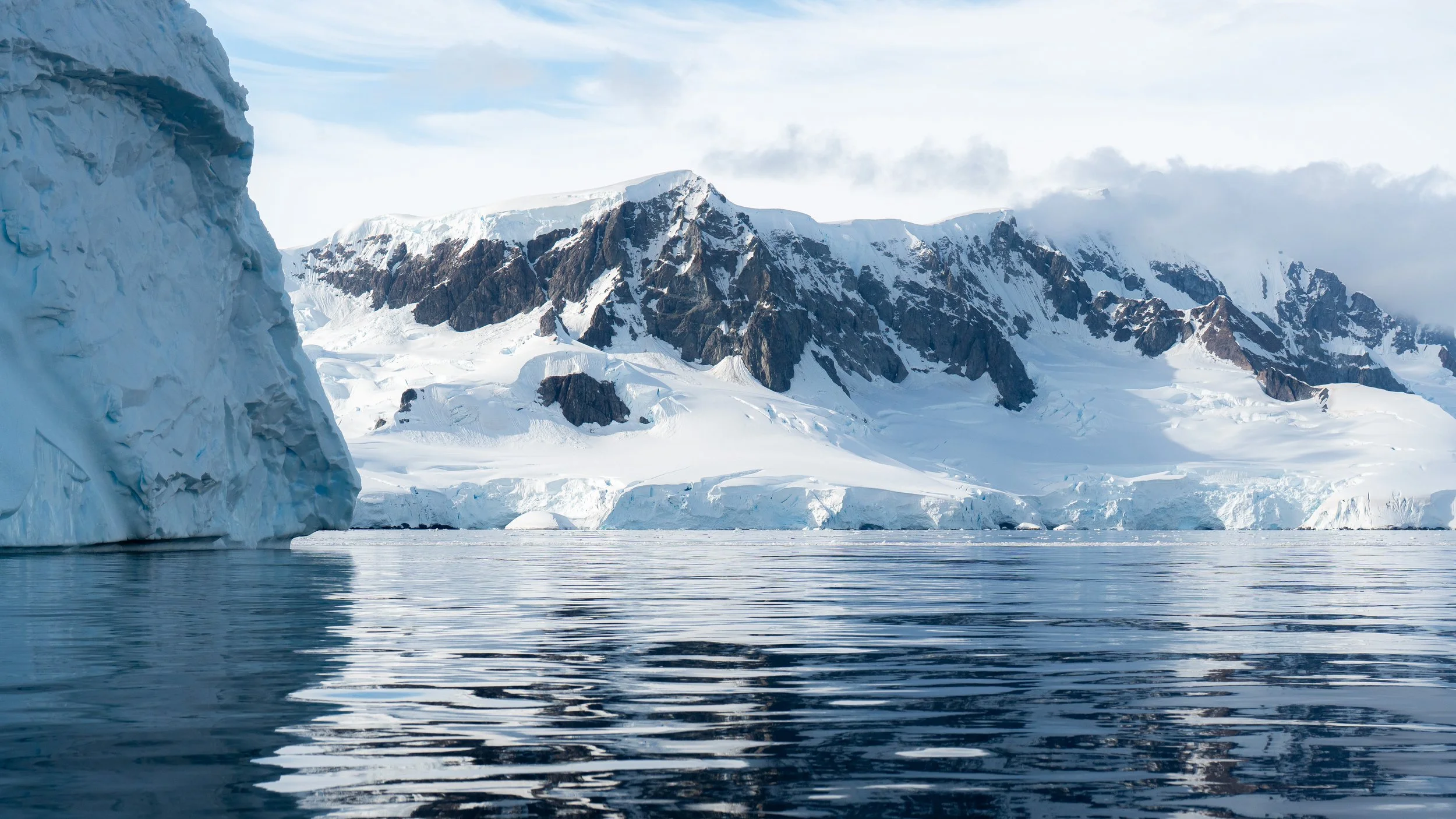 Snow-covered mountains and glaciers near calm ocean water, with a large ice formation on the left side and clouds in the sky.
