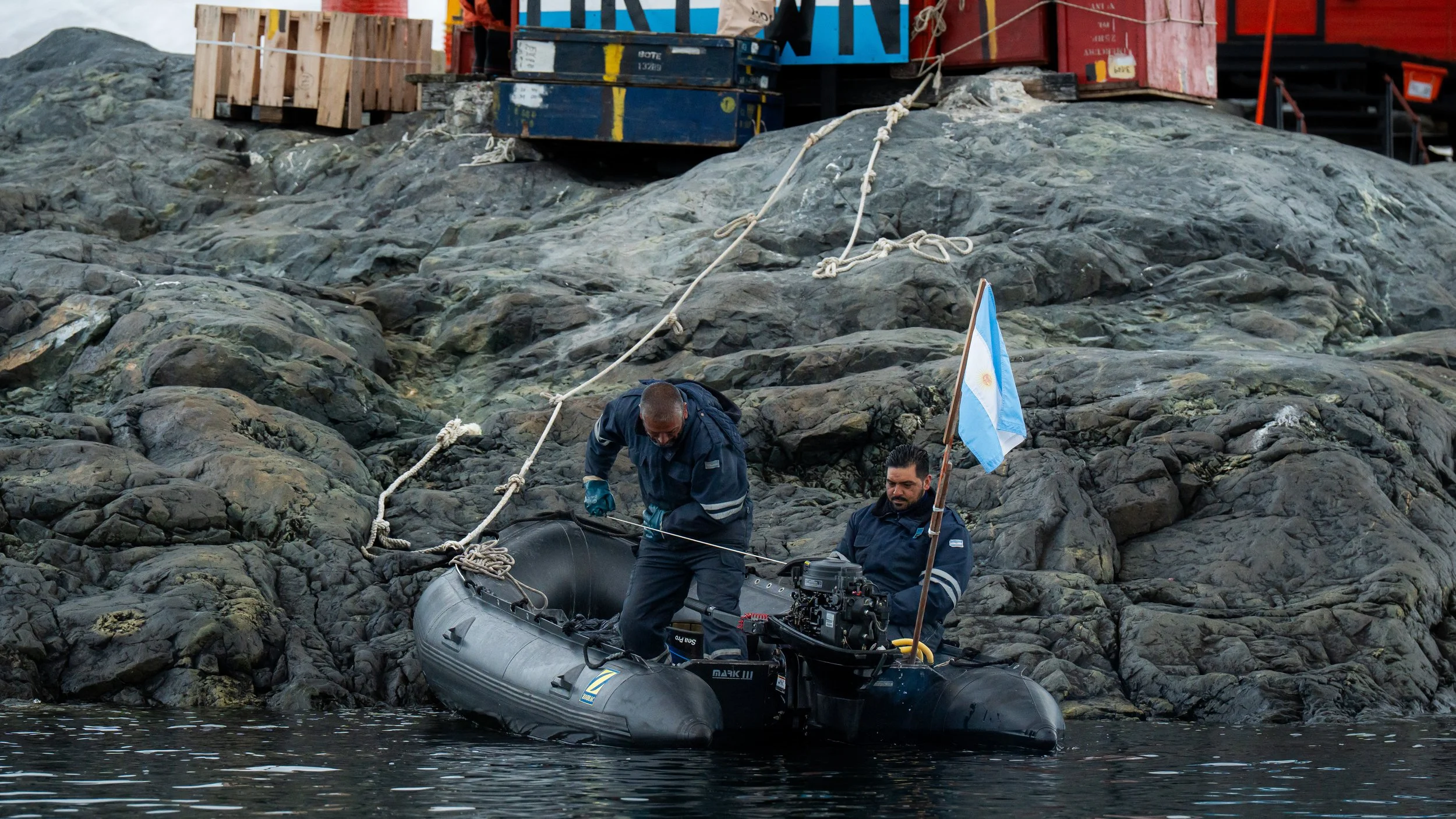 Two men in dark clothing are working on a gray inflatable boat near rocky shoreline, with one man holding onto the boat and the other adjusting equipment. One man is holding an Argentine flag. In the background, there are crates and a rope connected 