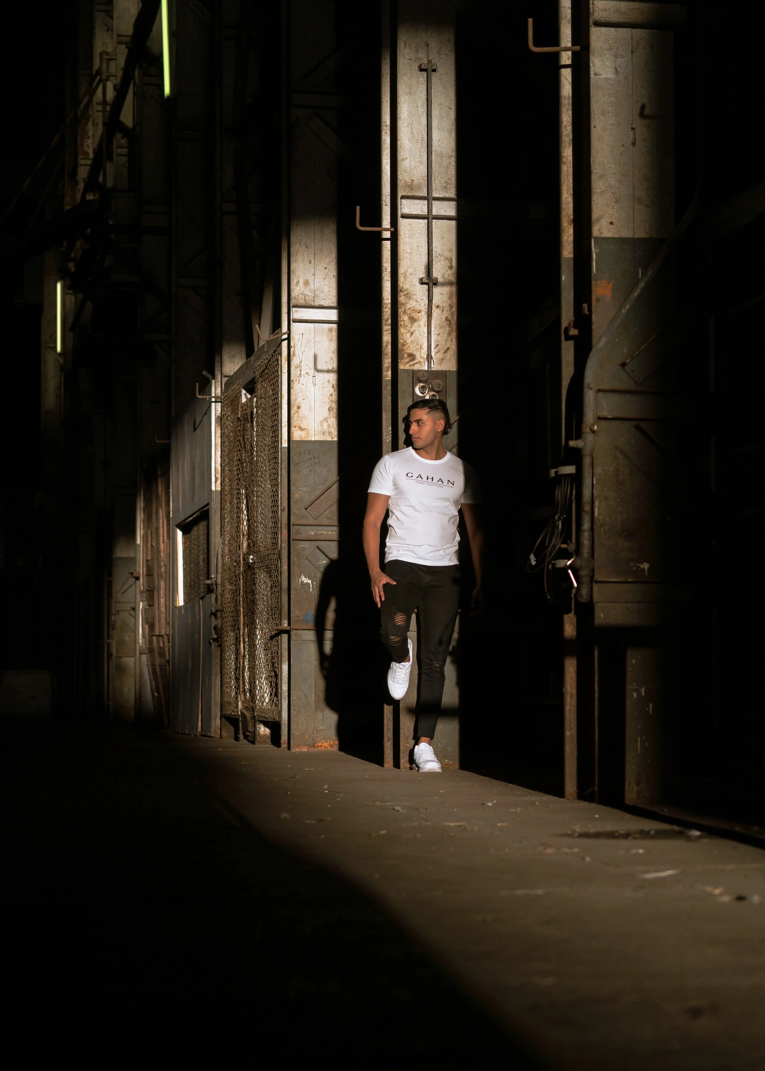 A young man with short dark hair, wearing a white Gahan T-shirt and ripped black jeans, runs through an industrial alleyway illuminated by partial sunlight, with metal gates and concrete walls.