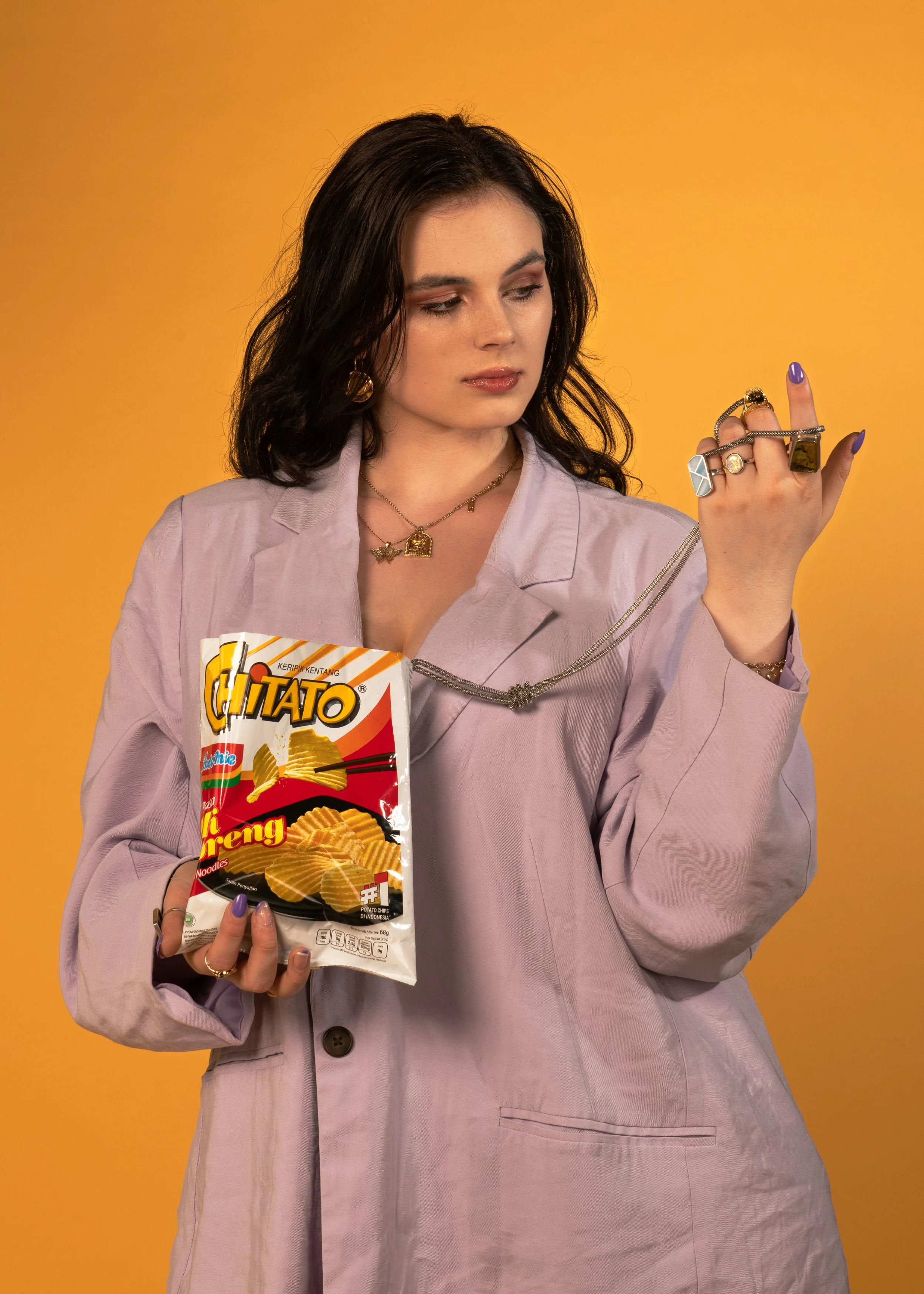 A woman with black wavy hair and gold earrings holds a bag of potato chips in her left hand and looks at her rings on her right hand, which are adorned with various jewelry pieces, against a plain yellow background.