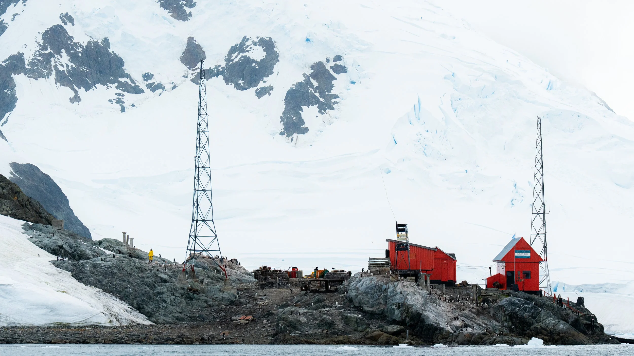 Base camp with red buildings, communication towers, and equipment on rocky, snowy terrain at the foot of a large icy mountain.
