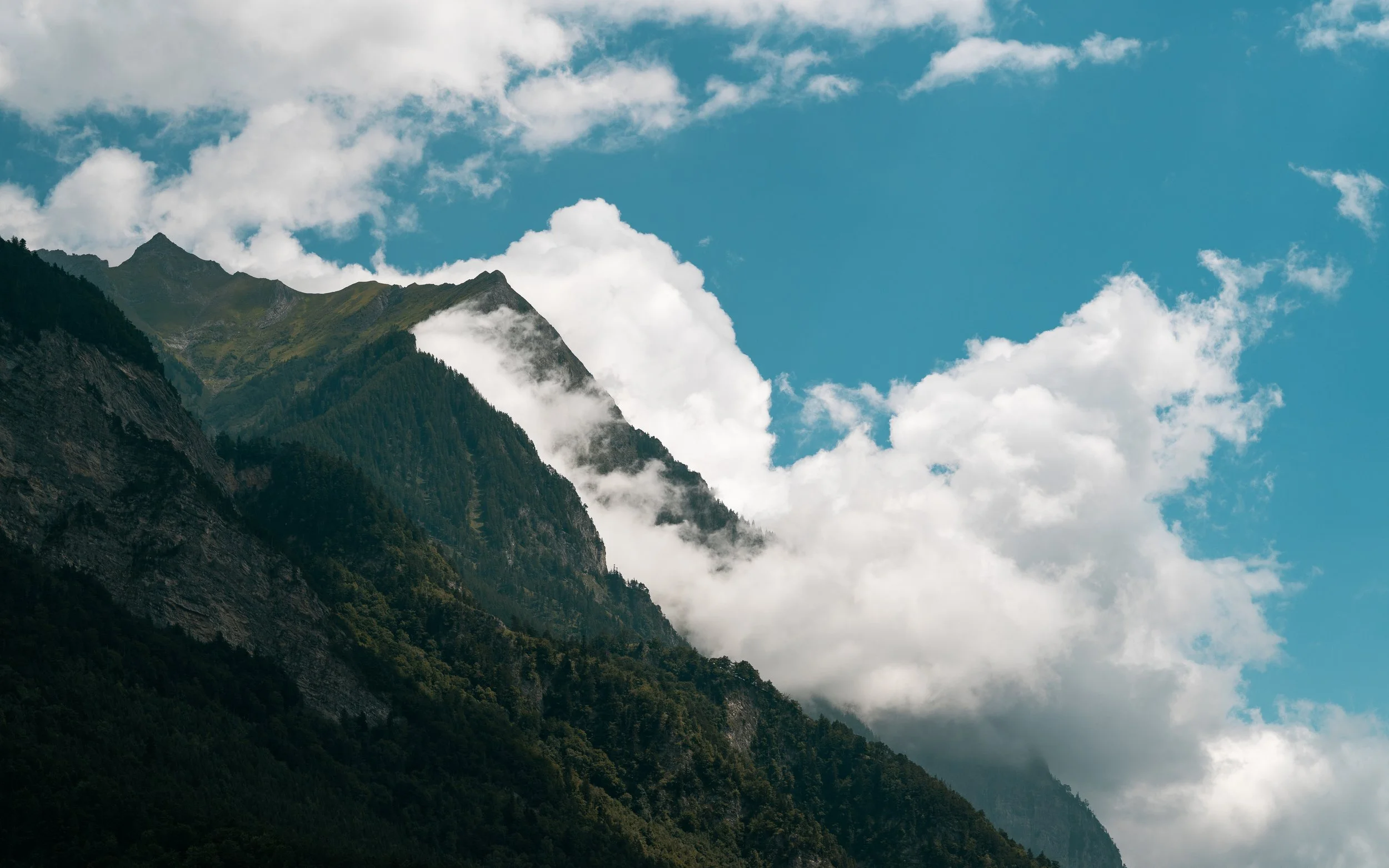 Mountains with green slopes, clouds surrounding peaks, and a blue sky with white clouds in the background.
