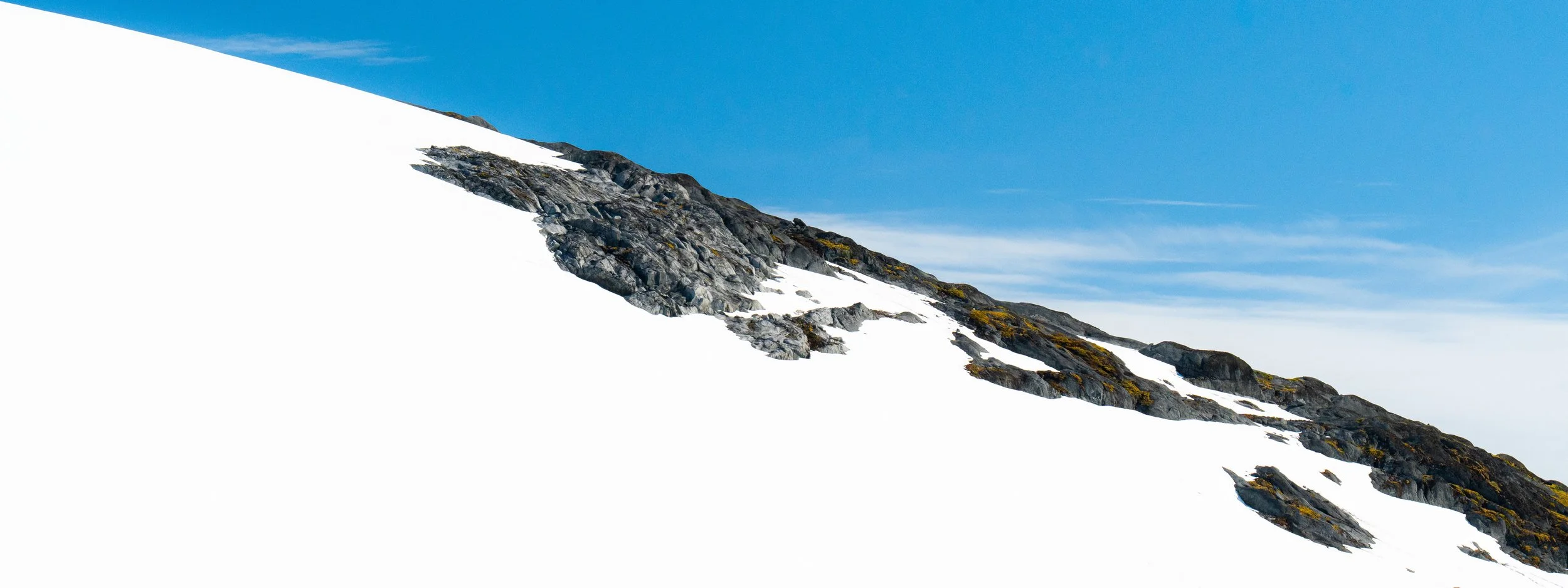 Snow-covered mountain slope with rocky patches, under a blue sky.