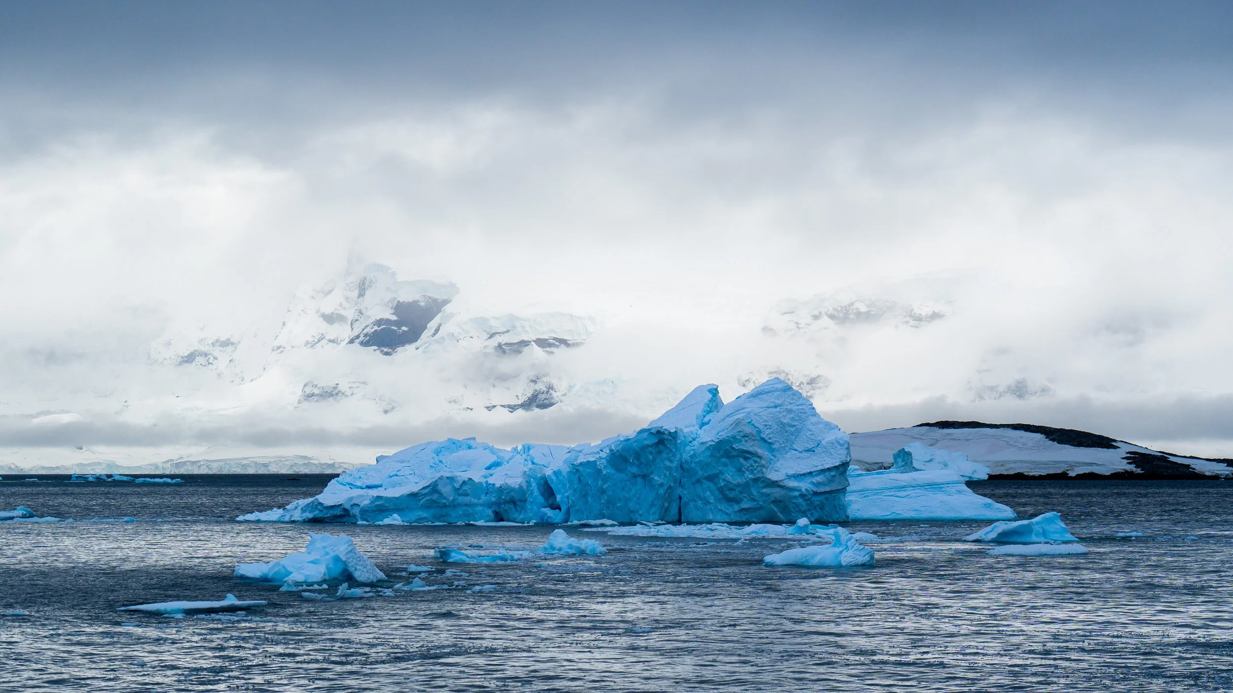 Icebergs floating in the ocean with snow-covered mountains and cloudy sky in the background