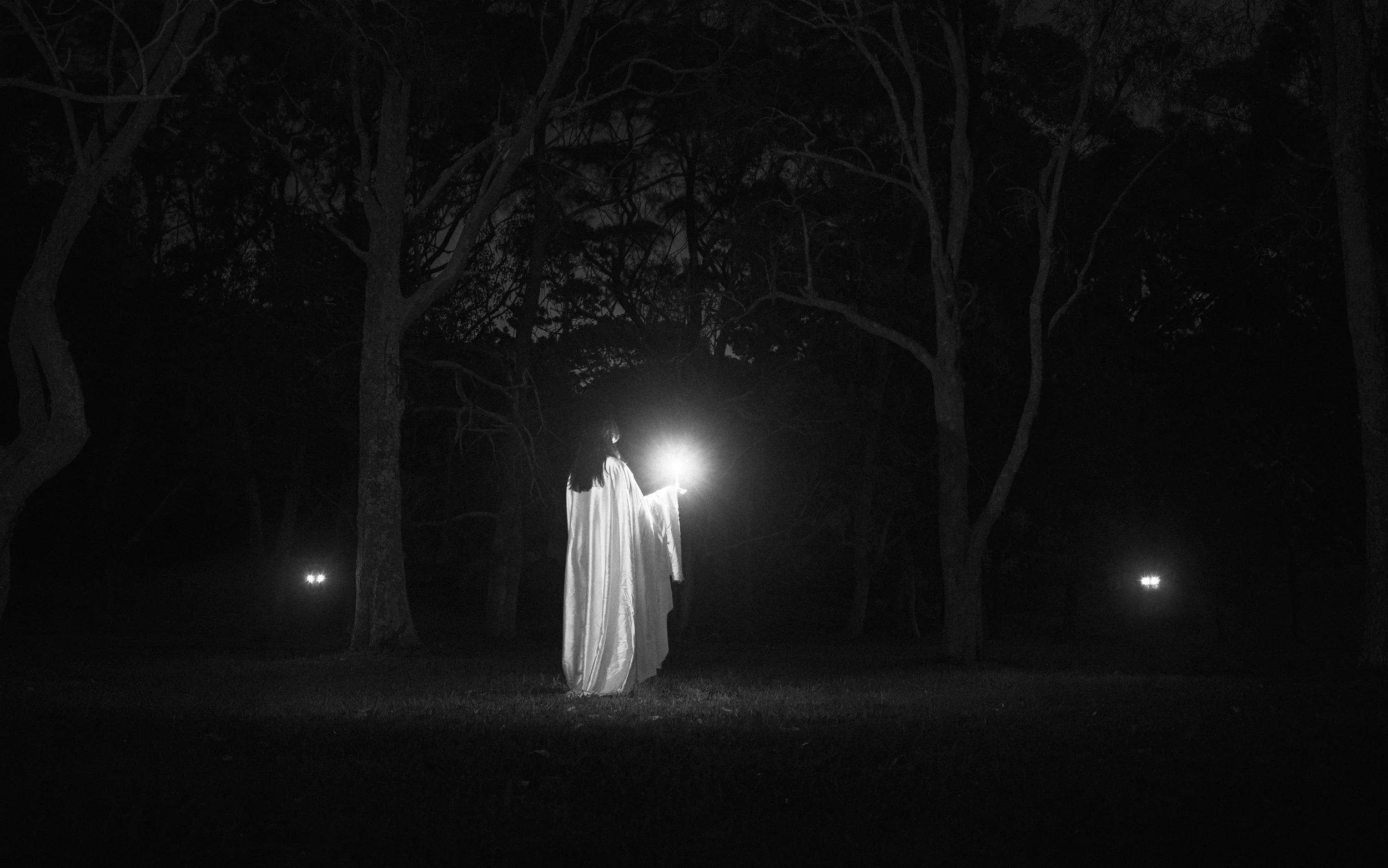A person wearing a white robe stands in a dark outdoor setting at night, holding a bright light source in their right hand that illuminates the surrounding trees. The scene appears mysterious and eerie with shadows cast by the trees.