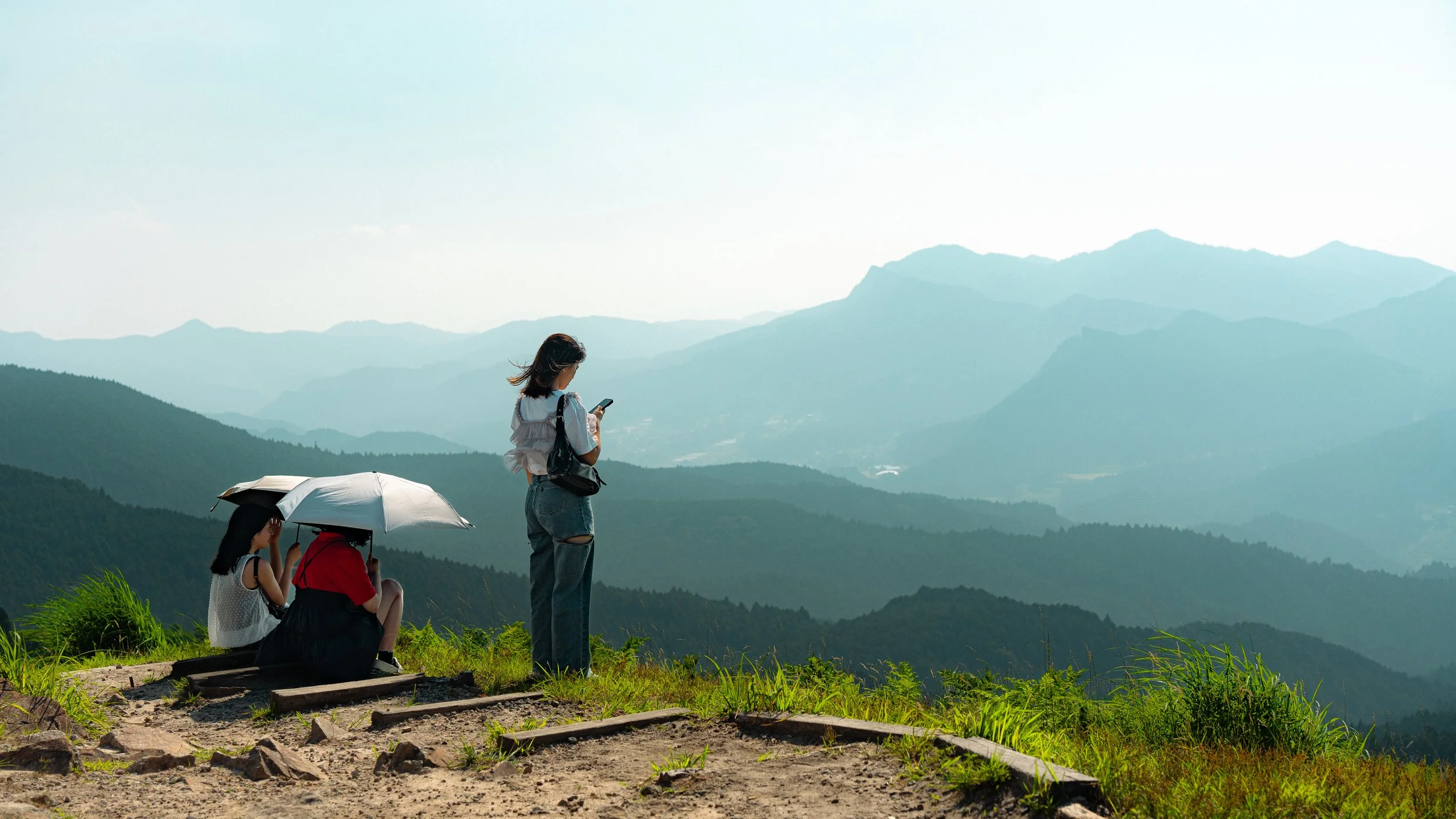 A woman standing on a grassy hill overlooking mountains, using a smartphone, while three people sit behind her under an umbrella.