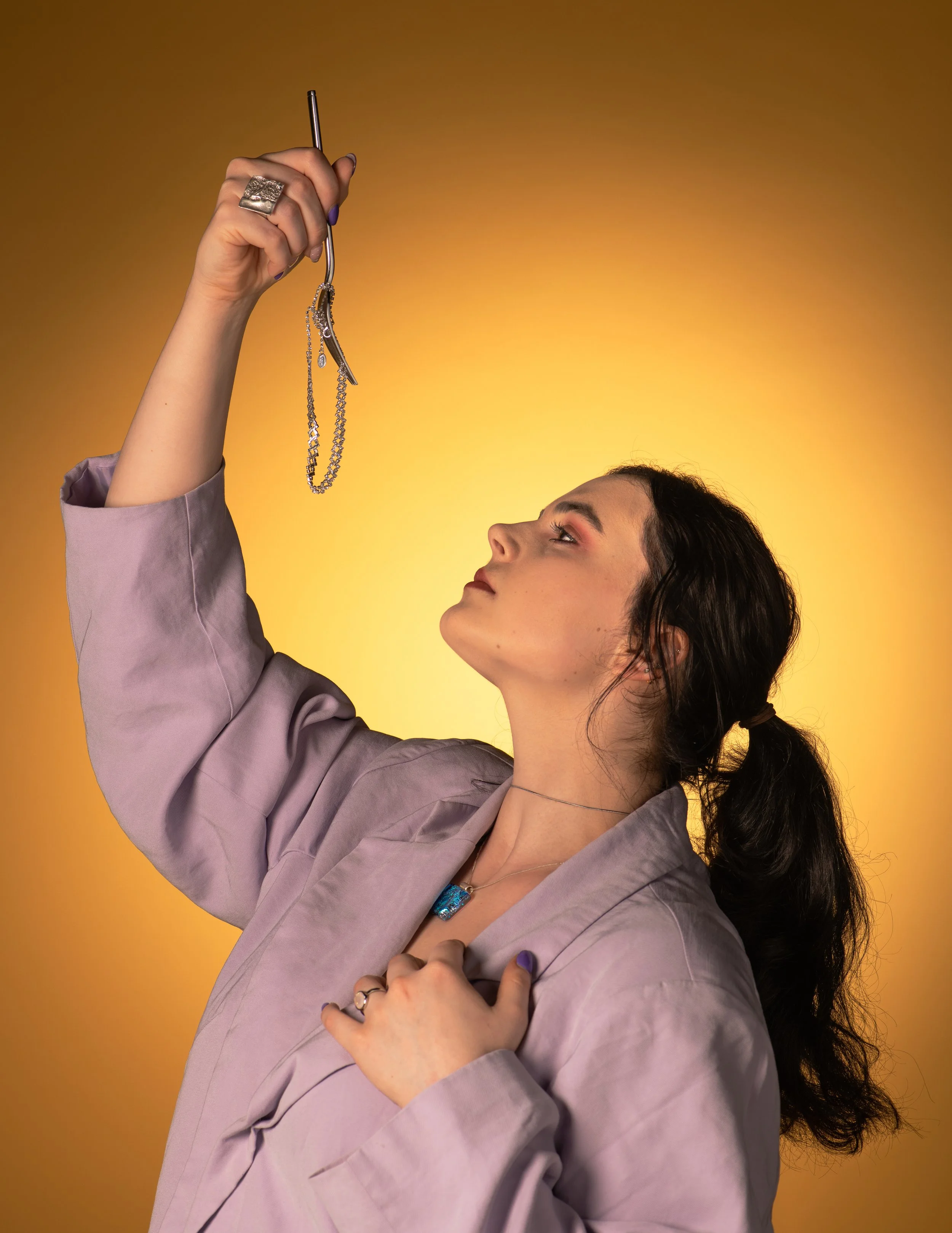 A woman with dark hair tied back, wearing a light purple jacket, gazing upwards at her hand that is holding a jewelry tool with a silver chain necklace, silver earrings, and rings, against a yellow gradient background.