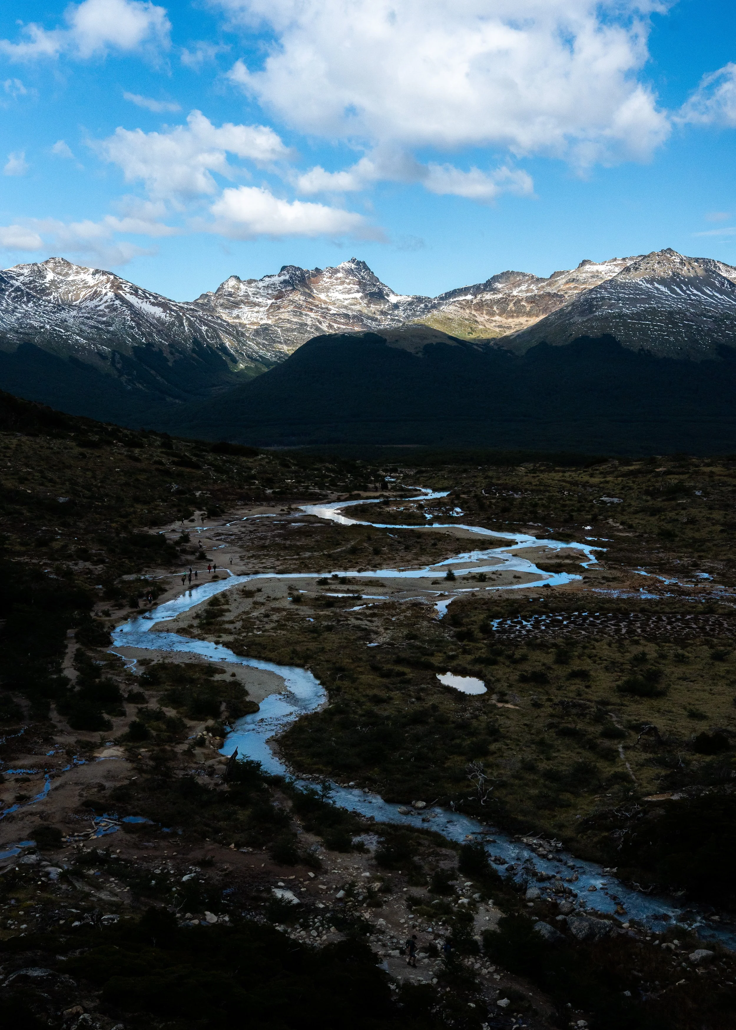 Scenic view of snow-capped mountains under a partly cloudy sky with a winding stream flowing through a dark, rocky terrain in the foreground. A few hikers are seen walking along the trail near the stream.