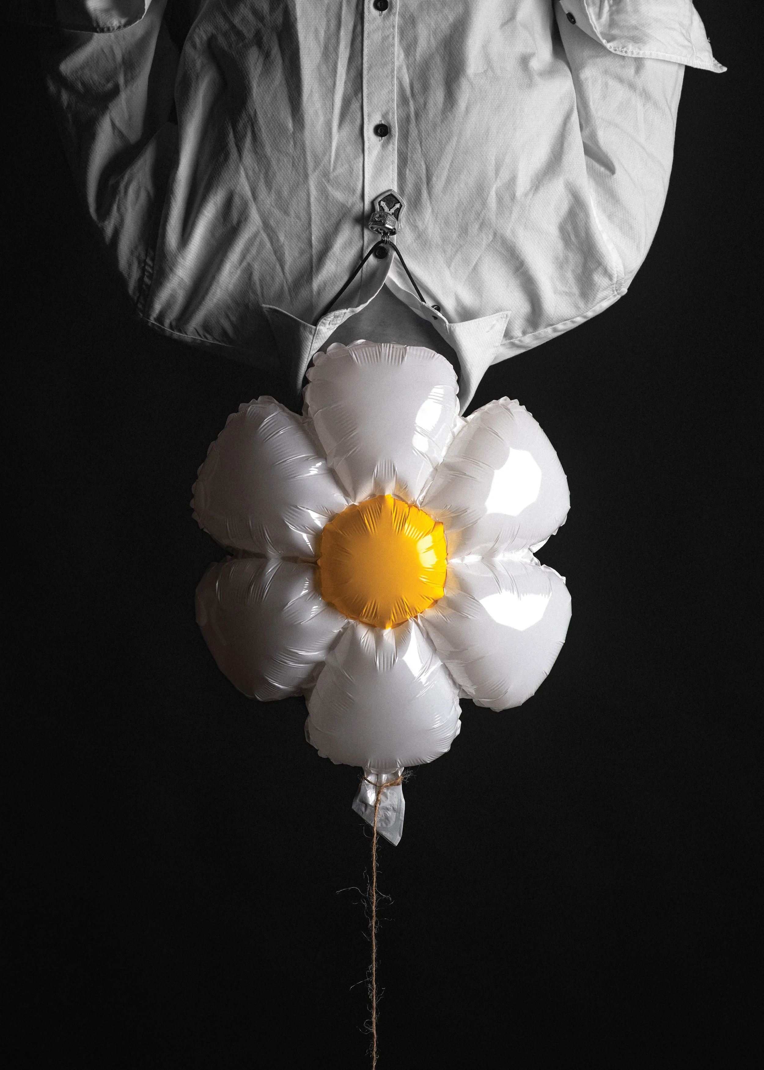 A bouquet of white balloons shaped like a flower with a yellow center, attached to a string, against a black background. Part of a person's upper body dressed in a light-colored shirt and jacket is visible above the balloons.