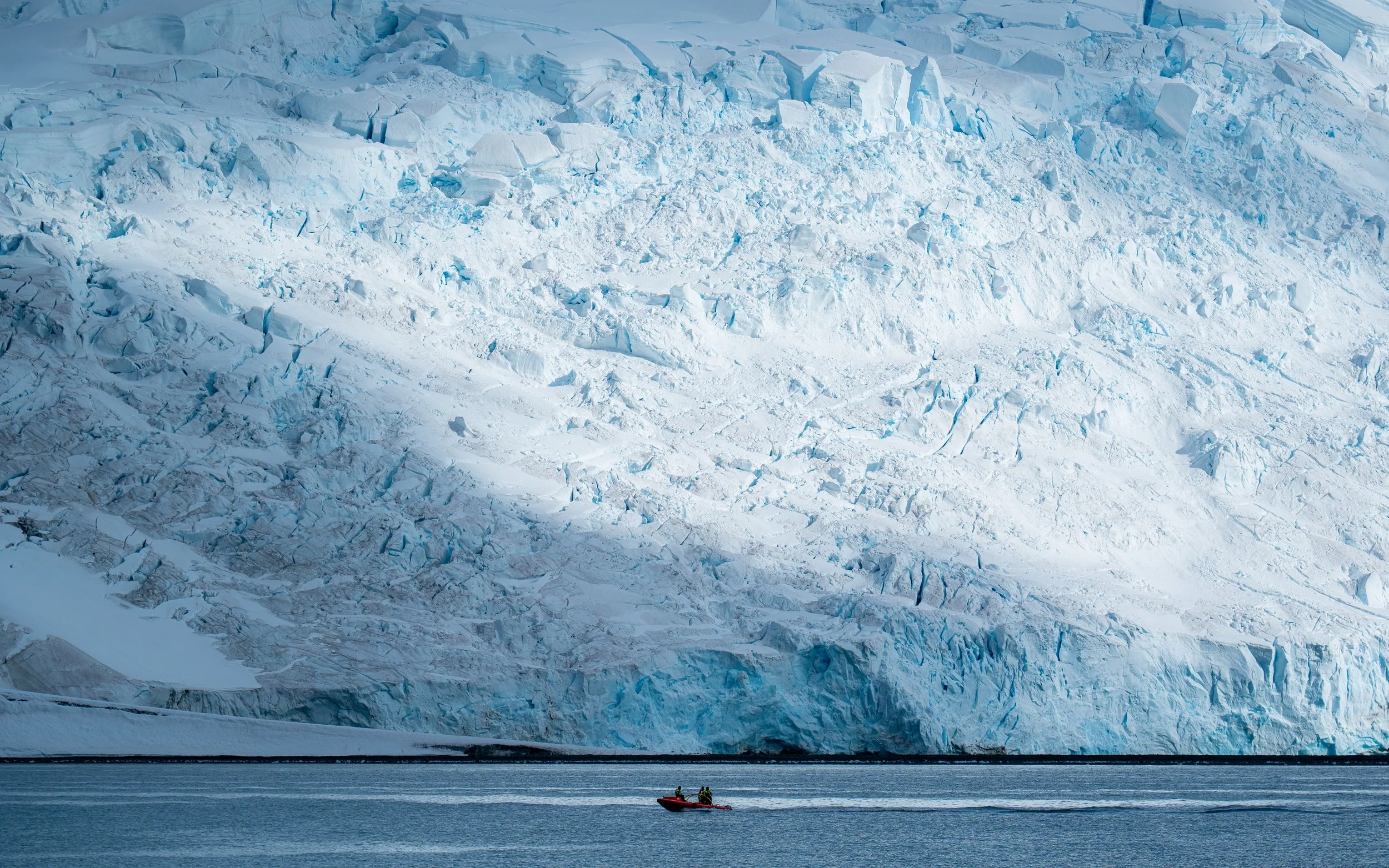 Two people in a red boat near a large icy glacier with a multicolored icefall in the background.
