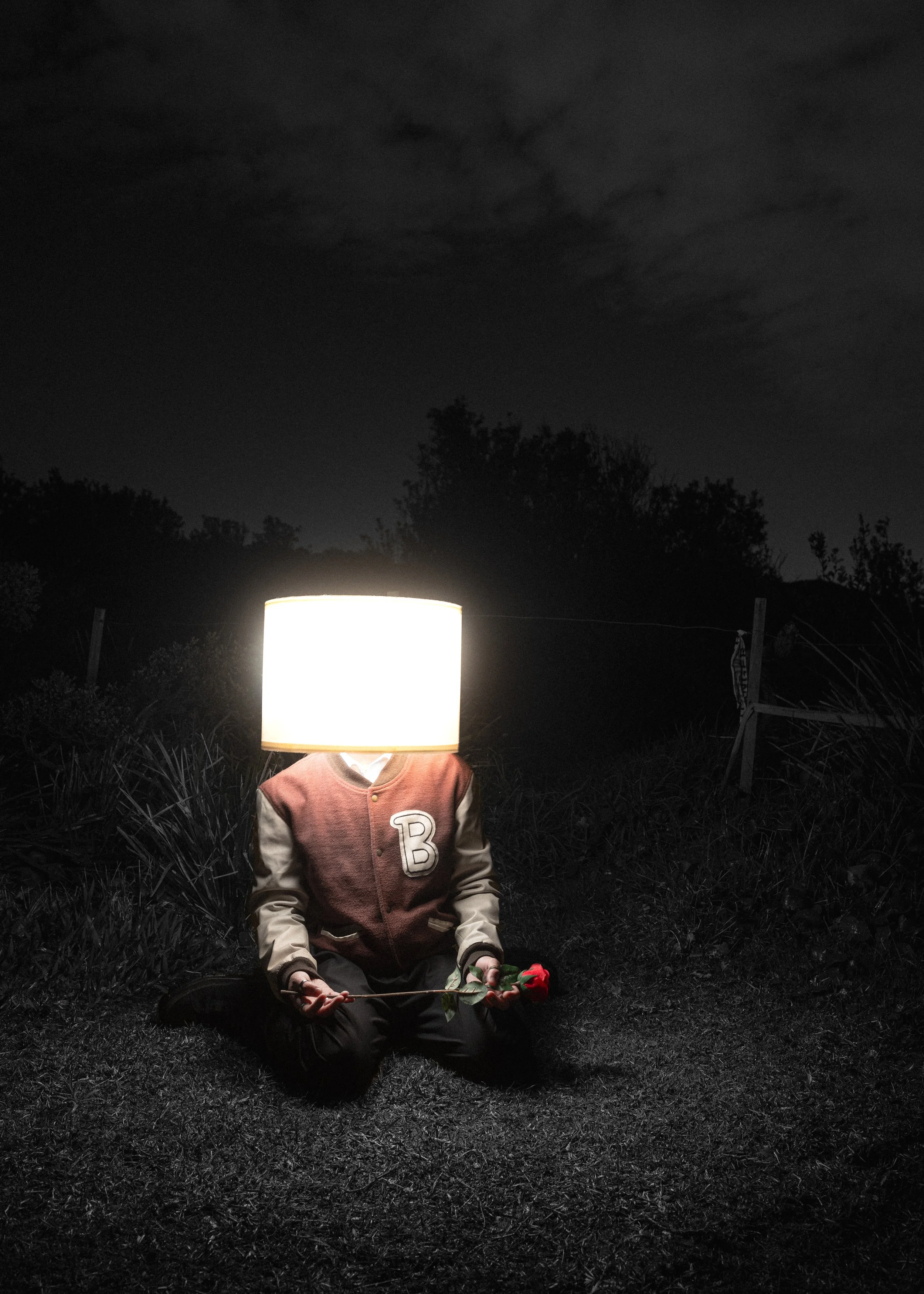 A person sitting on the ground at night with a lamp on their head, wearing a varsity jacket with the letter 'B' on it, holding a rose, with trees and a weather vane in the background.