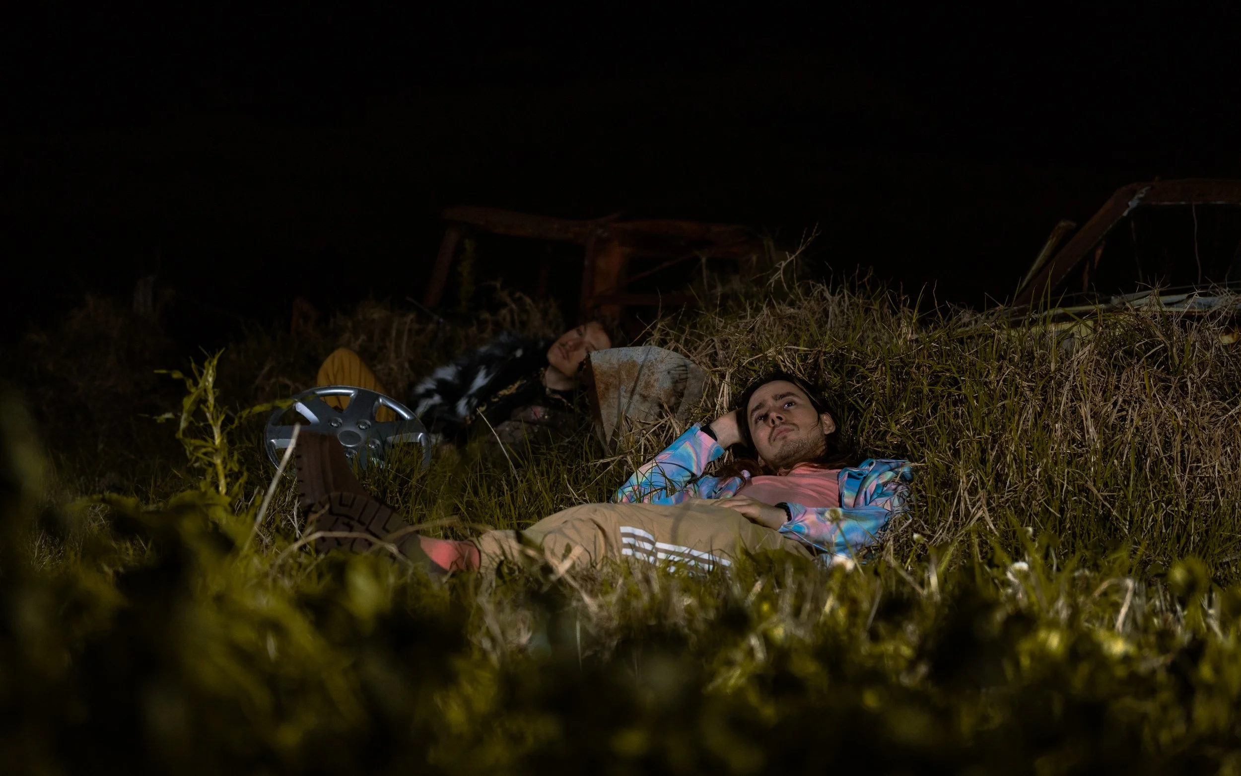 Two young adults lying in tall grass at night, looking upwards. One is a woman with long dark hair wearing a colorful jacket, and the other is a man with short dark hair wearing a dark jacket. The scene appears to be outdoors with some old, rusty mac