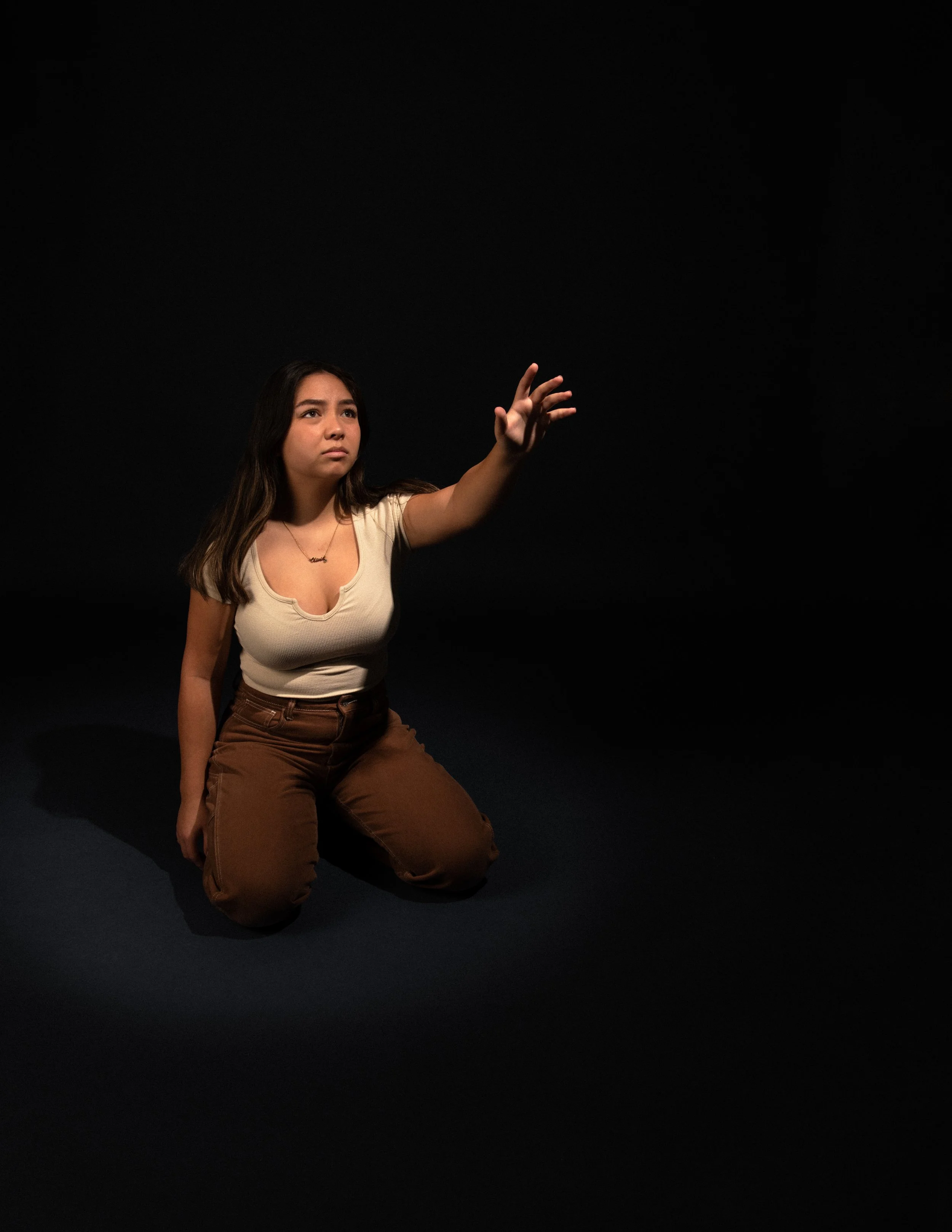 A young woman kneeling on the floor in front of a black background, reaching out with her right hand while looking slightly upward with a serious expression.