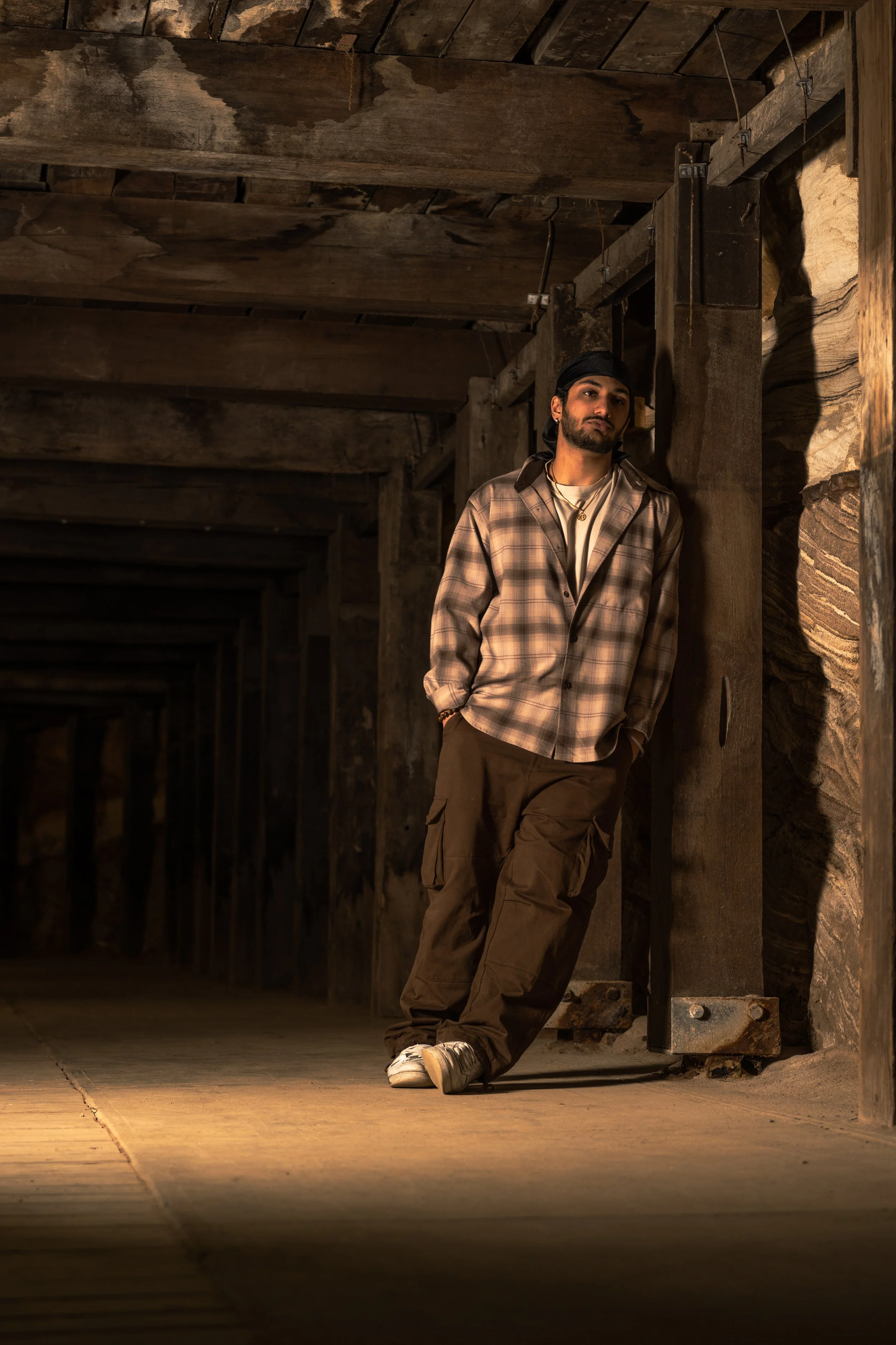 A young man with a beard and mustache, wearing a plaid shirt, cargo pants, sneakers, and a black bandana, leaning against a wooden post in an underground tunnel with wooden beams and stone walls.