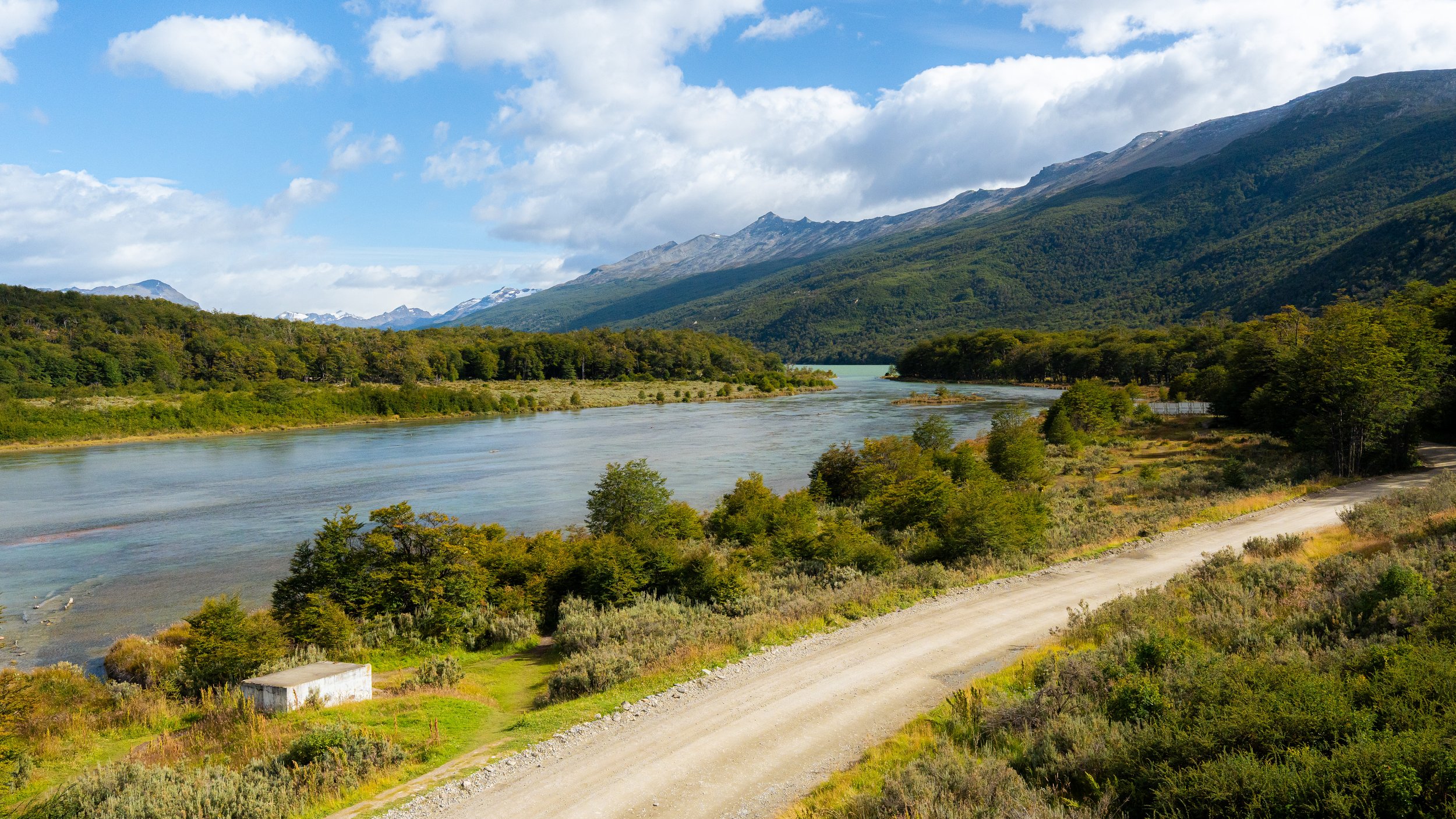 Scenic landscape of a river flowing through green forests with mountains in the background and a partly cloudy sky.