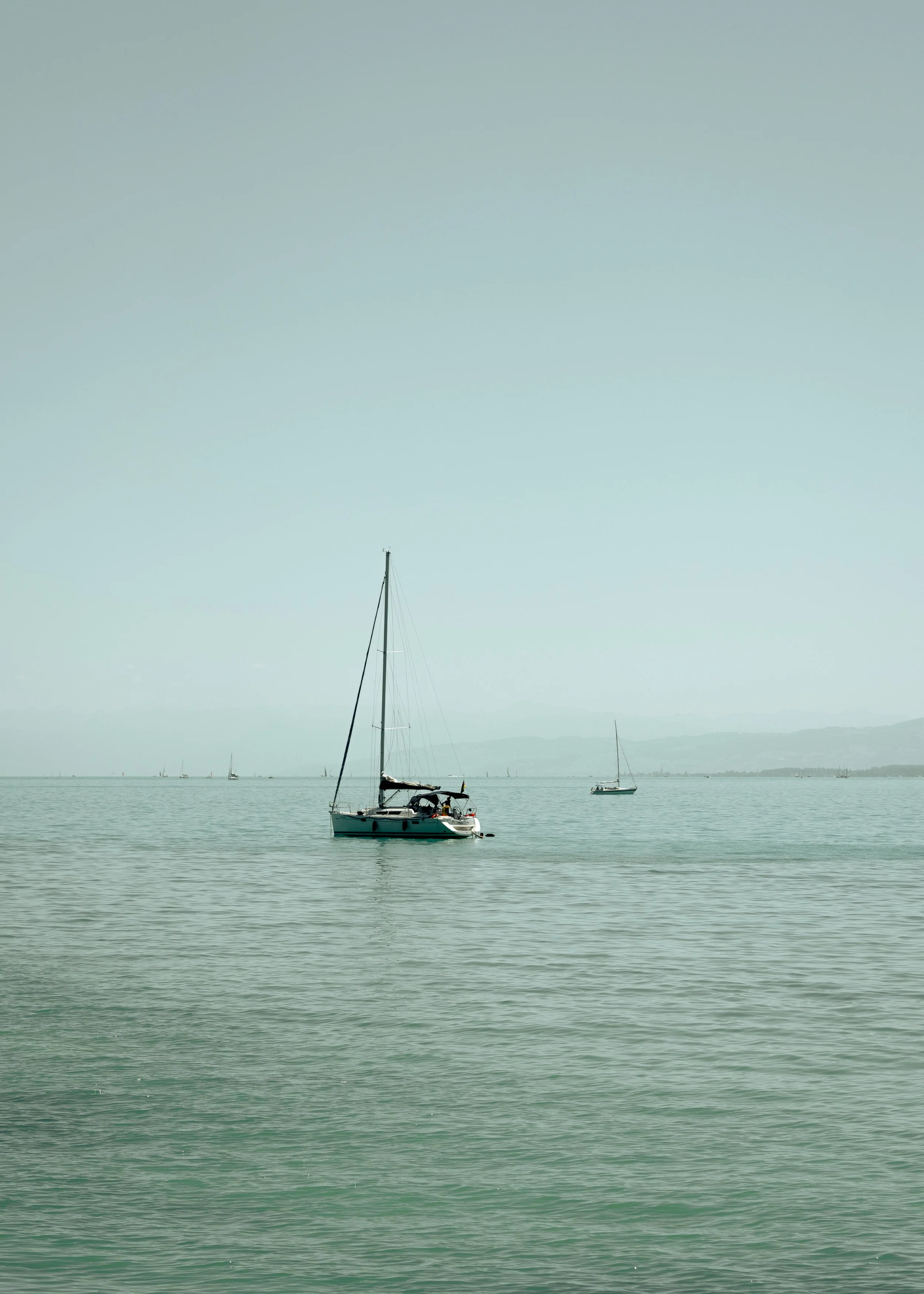 A sailboat on calm water with other sailboats in the distance, mountains in the background, under a clear sky.