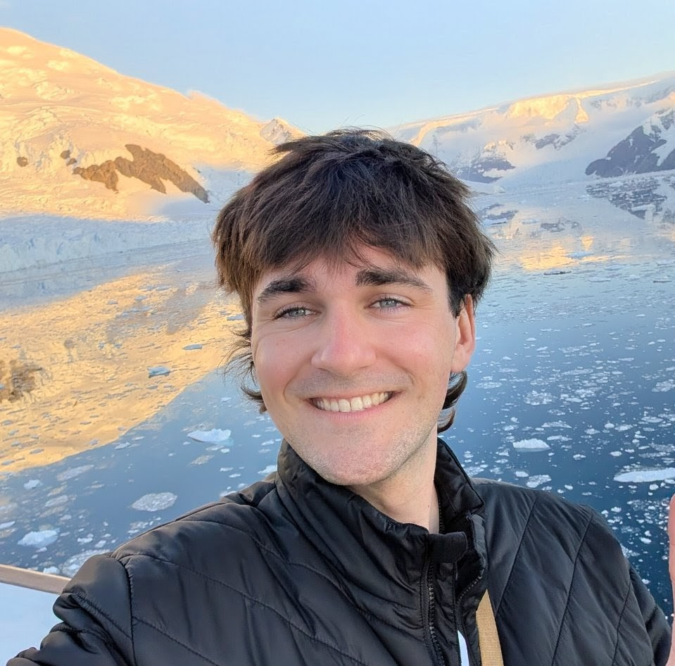Smiling young man with dark hair in an outdoor icy landscape with snow-covered mountains and icy water in the background.