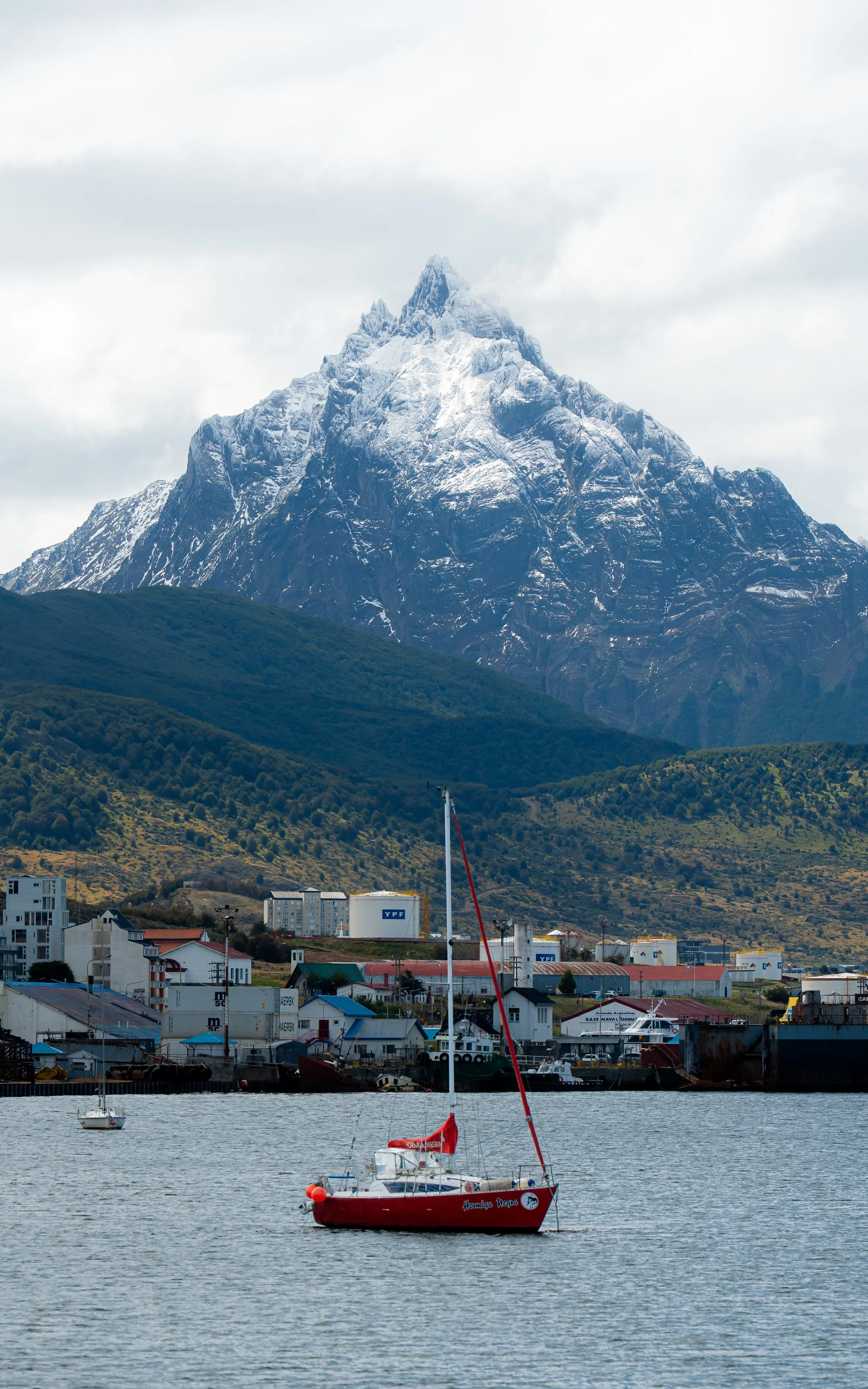 A red sailboat floating on calm water with a mountain in the background and a small town at the water's edge.