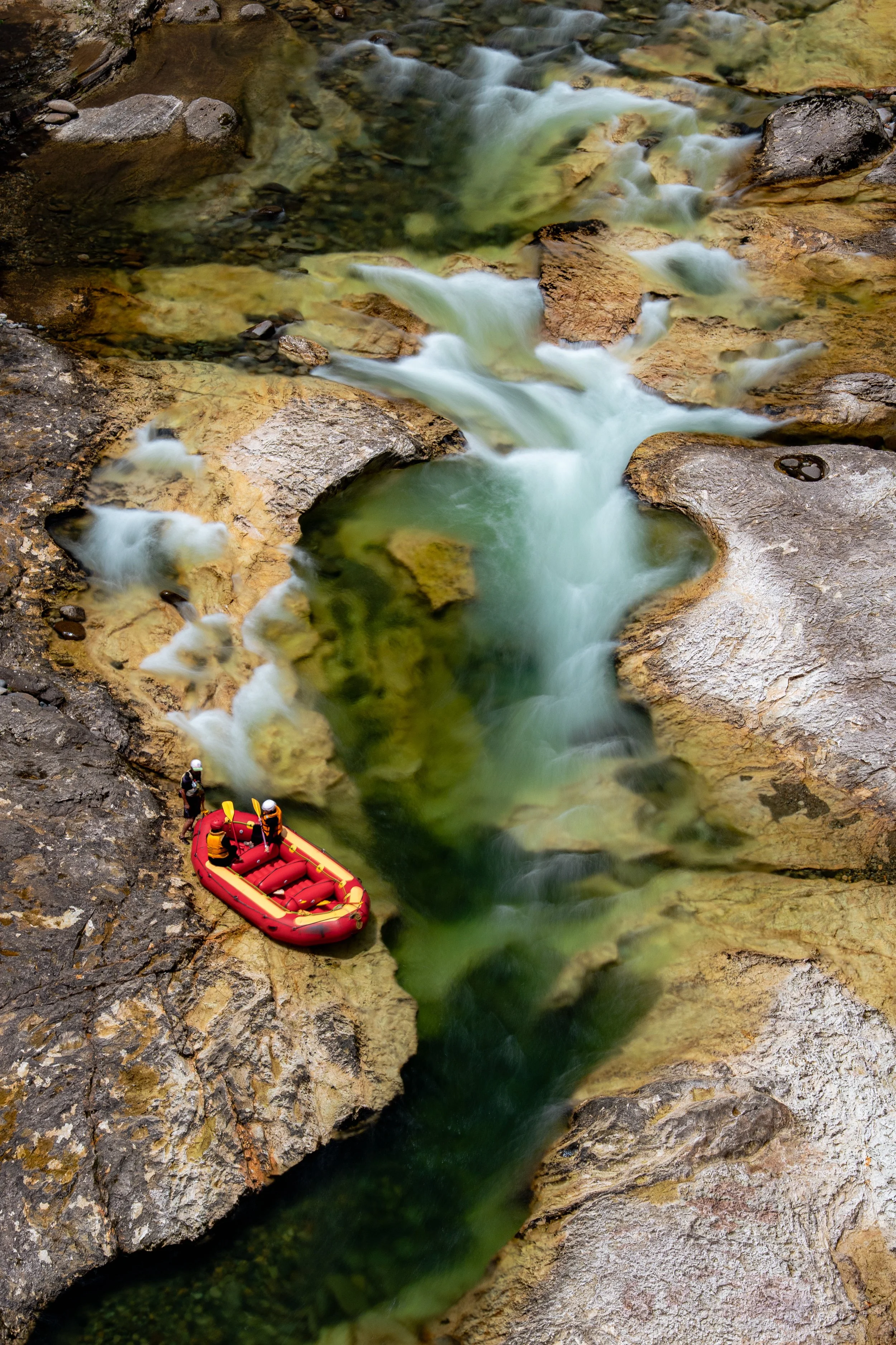 Five people in helmets and life jackets on a red inflatable raft navigating through a narrow rocky canyon with rushing water.