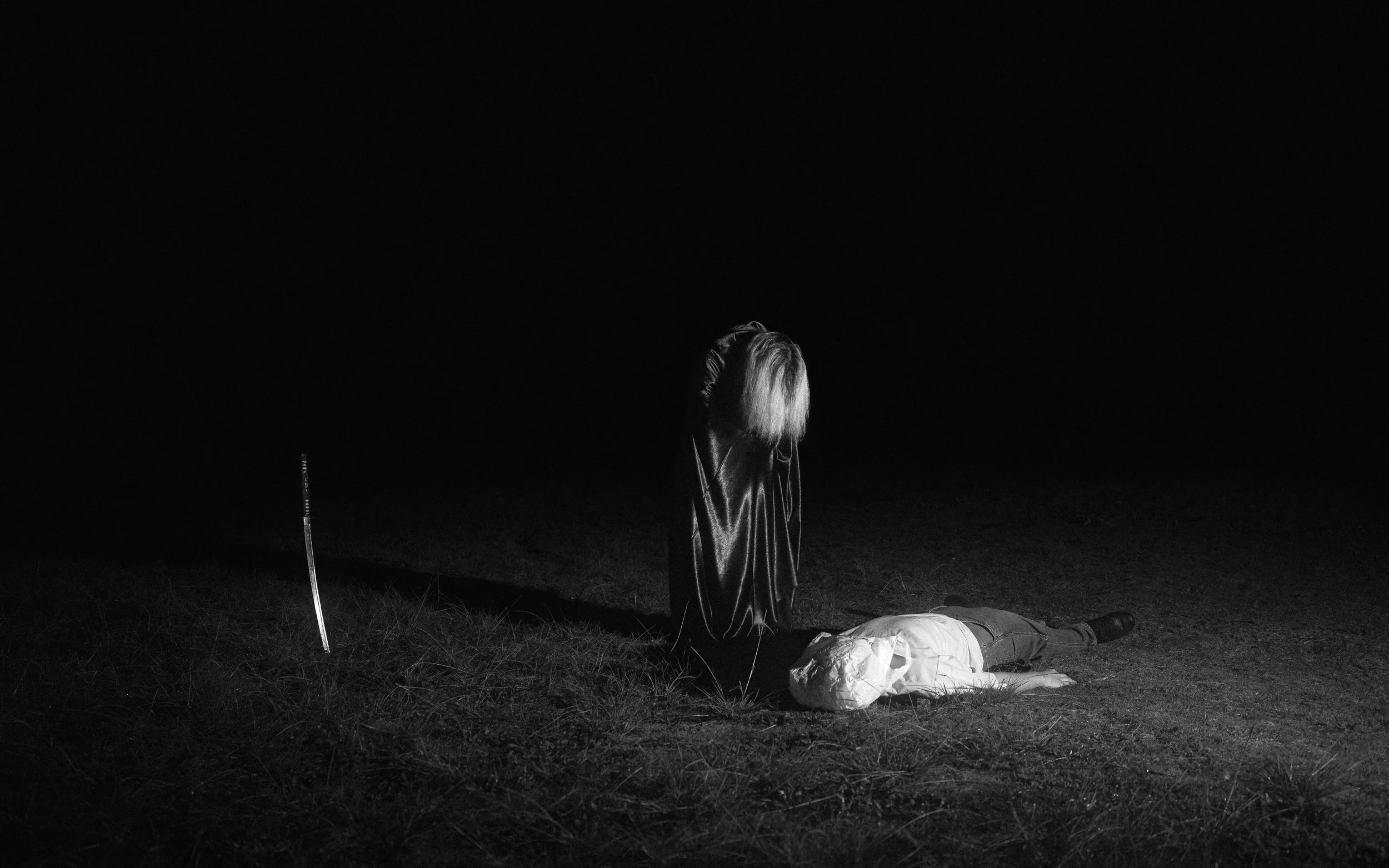A woman with long hair reclining on the ground at night, with camping gear including a sleeping bag and a small tent nearby. The scene is dark with minimal lighting, creating a quiet, contemplative atmosphere.