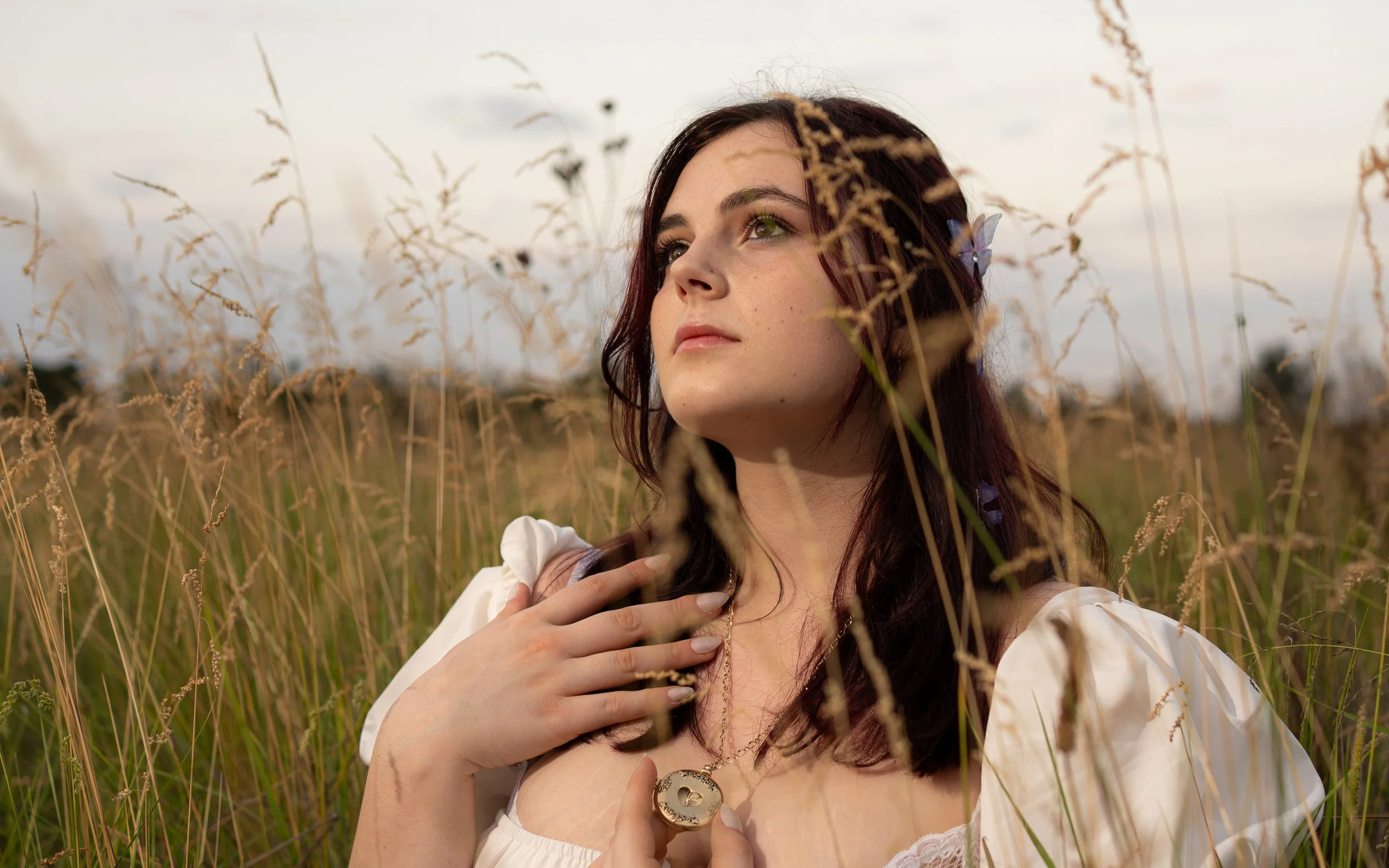 A woman with dark hair and green eyes stands in a field of tall, dry grass. She is wearing a white dress with puffed sleeves, a gold heart-shaped necklace, and has a butterfly on her hair. She is touching her chest with one hand and looking off into 