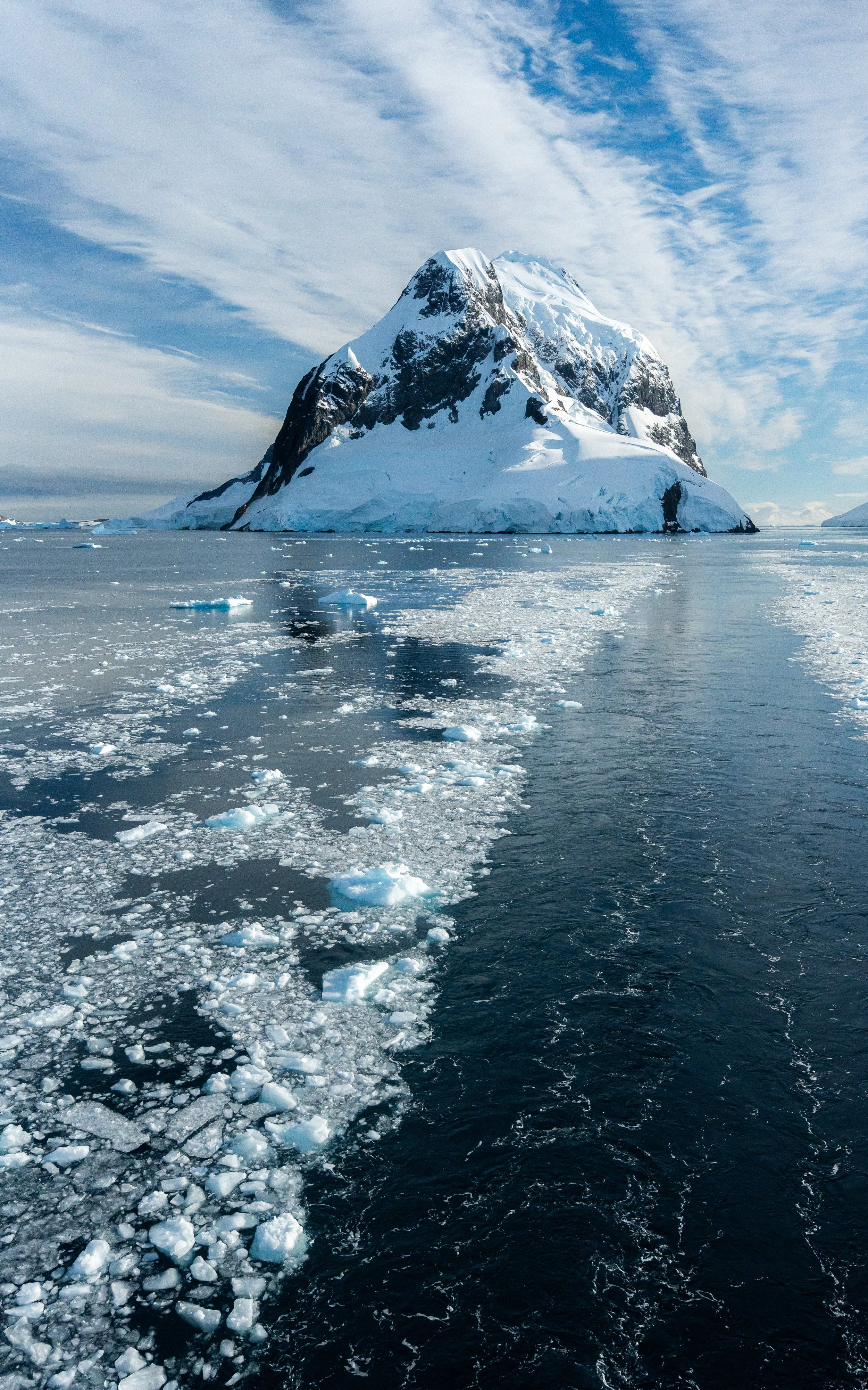 A large snow-capped mountain surrounded by ice and water, with a trail of floating ice chunks leading toward the mountain.